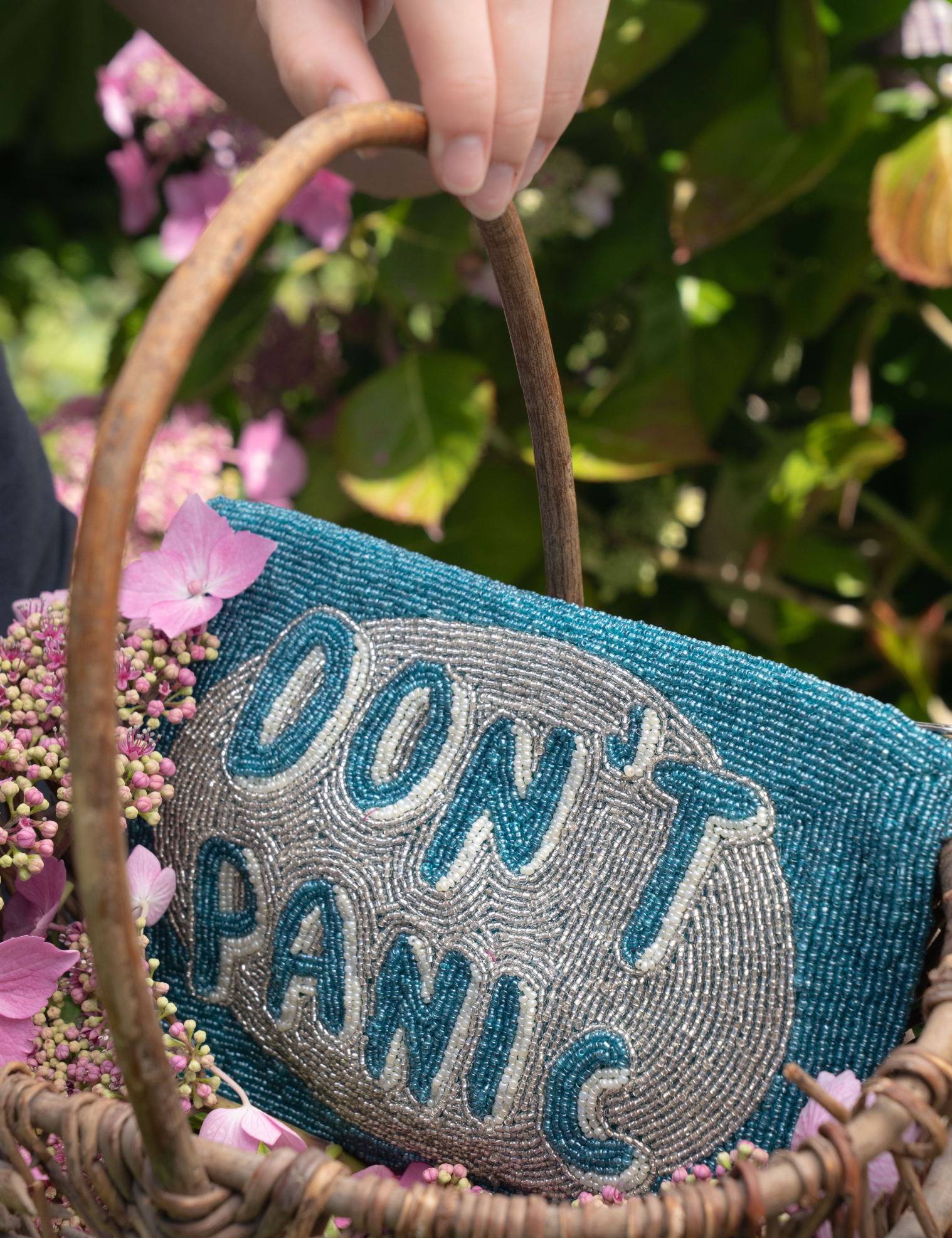 A close-up of a woven wicker basket with a blue and gray beaded clutch bag that reads 'DON'T PANIC' in raised letters. A hand is holding the basket handle, and pink flowers and green foliage are softly blurred in the background.