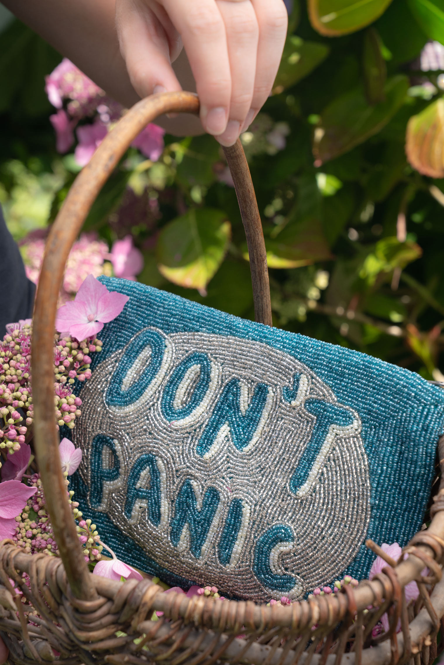 A close-up of a woven wicker basket with a blue and gray beaded clutch bag that reads 'DON'T PANIC' in raised letters. A hand is holding the basket handle, and pink flowers and green foliage are softly blurred in the background.