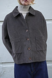 A person wearing a dark brown corduroy jacket over a cream ribbed turtleneck sweater and dark blue jeans, standing against a white textured wall.