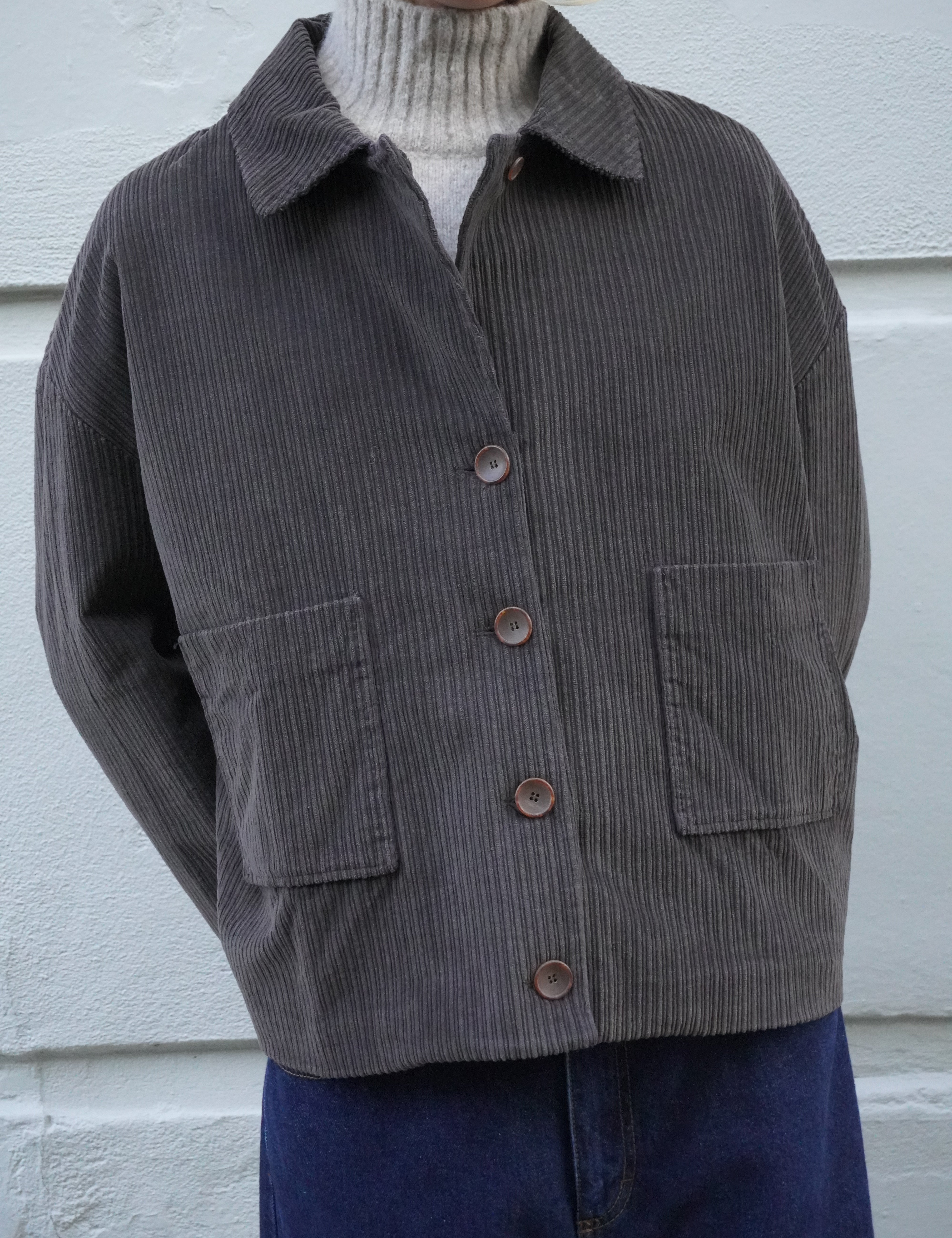 A person wearing a dark brown corduroy jacket over a cream ribbed turtleneck sweater and dark blue jeans, standing against a white textured wall.