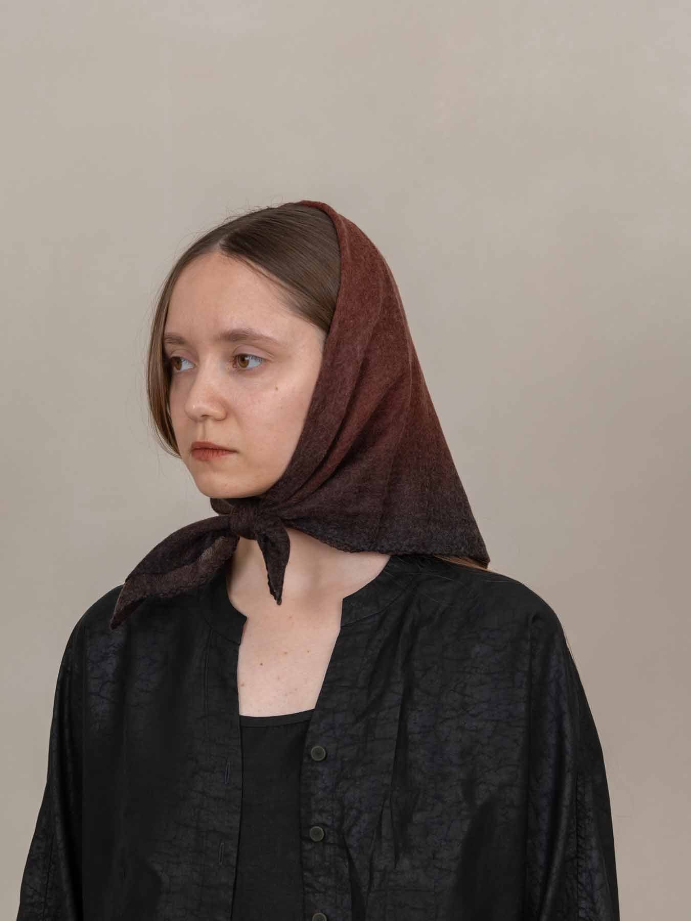 A woman wearing a dark brown knitted headscarf tied under her chin, paired with a black textured button-up shirt, set against a plain beige background.