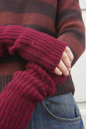 A close-up of a person wearing a chunky ribbed knit fingerless glove in deep burgundy over a brown and burgundy striped sweater, paired with blue jeans.