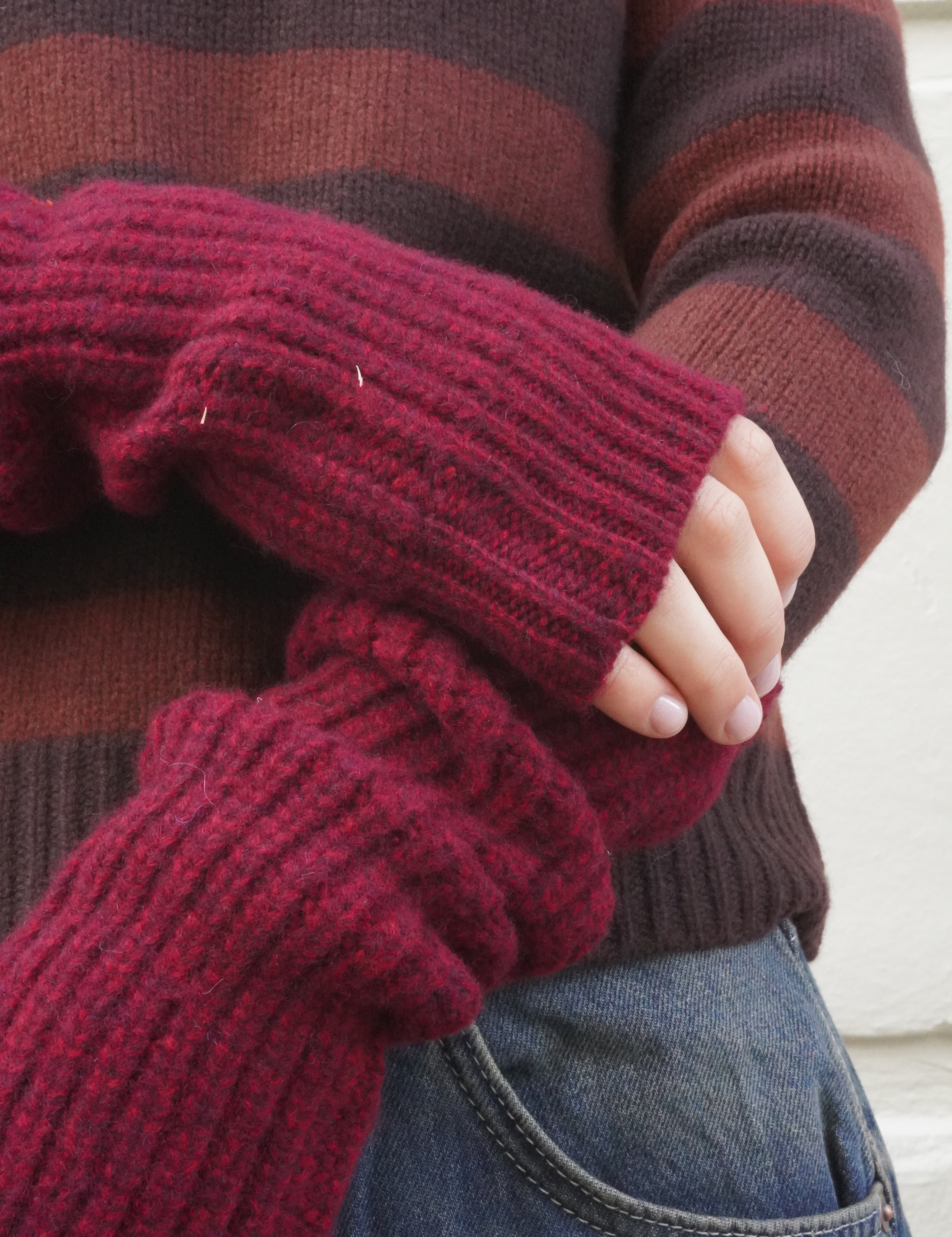A close-up of a person wearing a chunky ribbed knit fingerless glove in deep burgundy over a brown and burgundy striped sweater, paired with blue jeans.