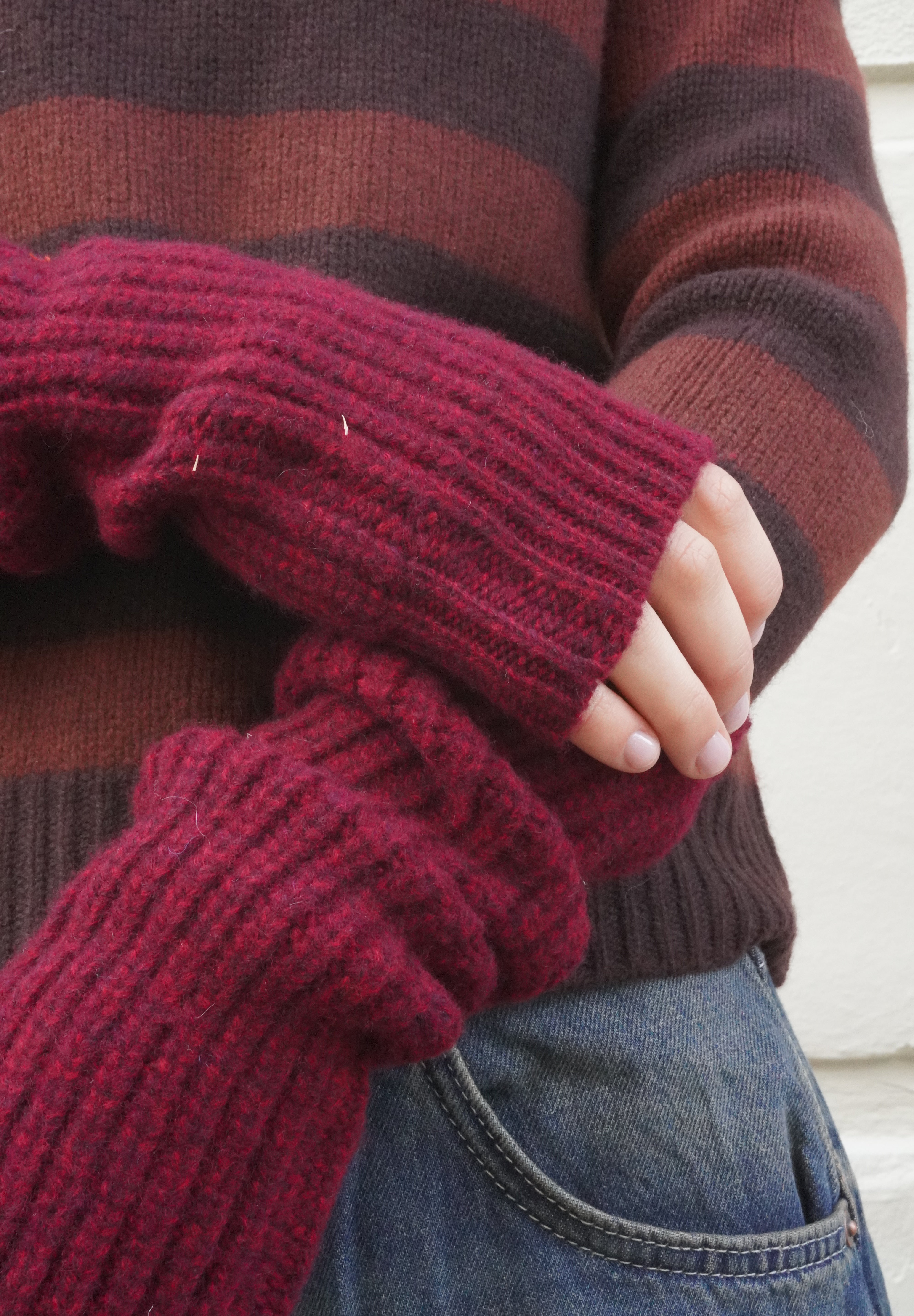 A close-up of a person wearing a chunky ribbed knit fingerless glove in deep burgundy over a brown and burgundy striped sweater, paired with blue jeans.