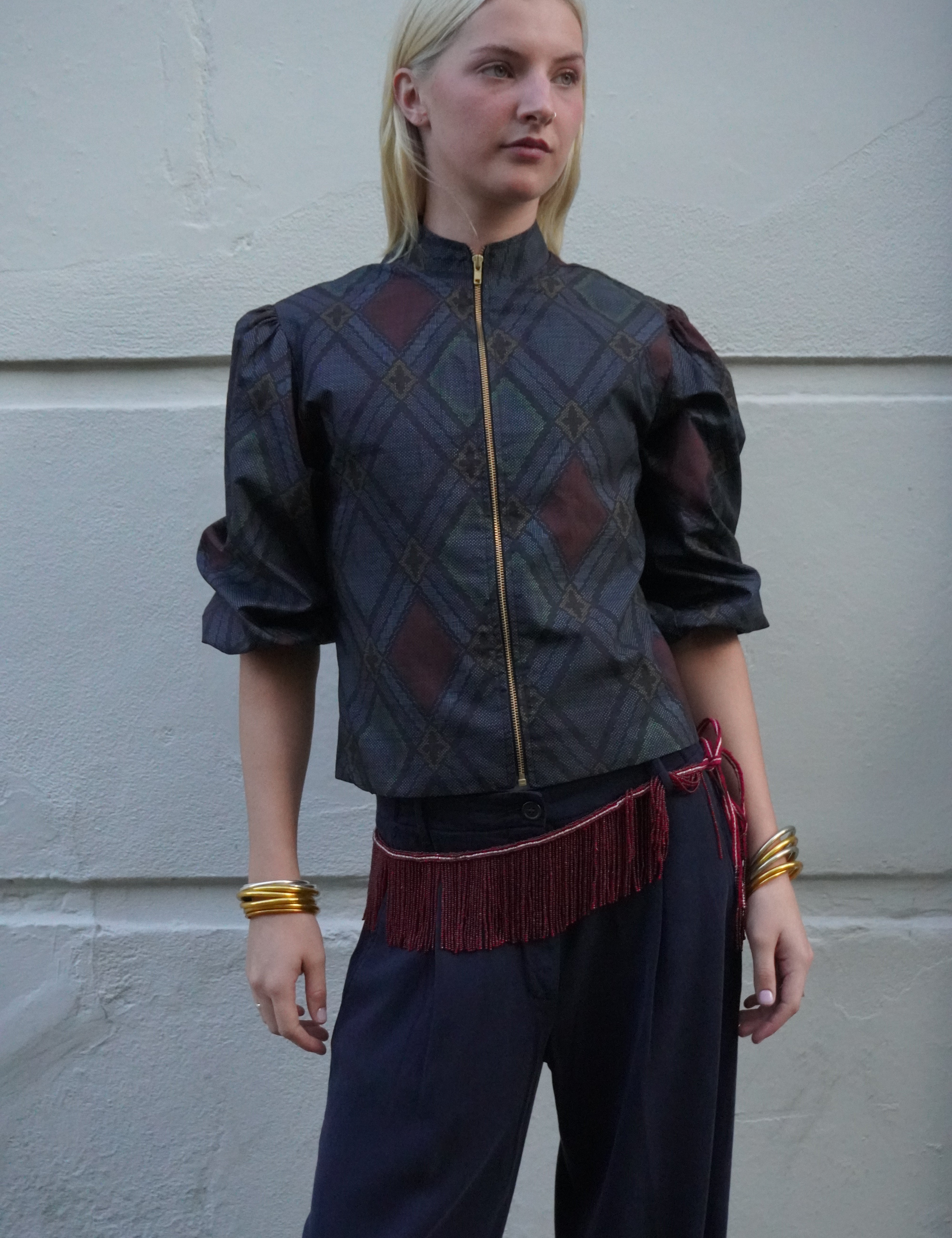 A woman stands against a light gray textured wall wearing a dark patterned cropped jacket with puff sleeves, a gold zipper, and a red fringe belt, paired with dark pants and gold bracelets.