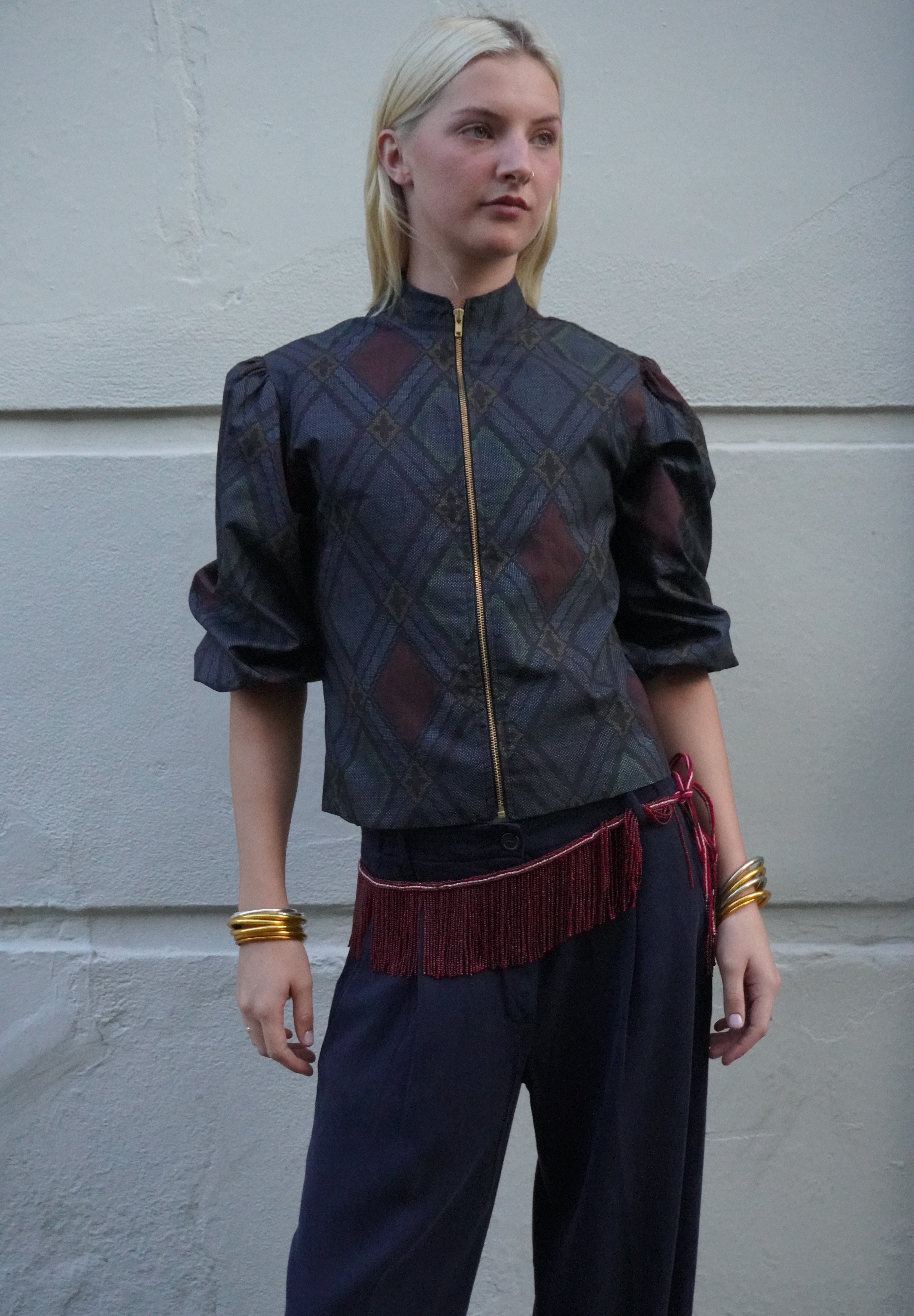 A woman stands against a light gray textured wall wearing a dark patterned cropped jacket with puff sleeves, a gold zipper, and a red fringe belt, paired with dark pants and gold bracelets.