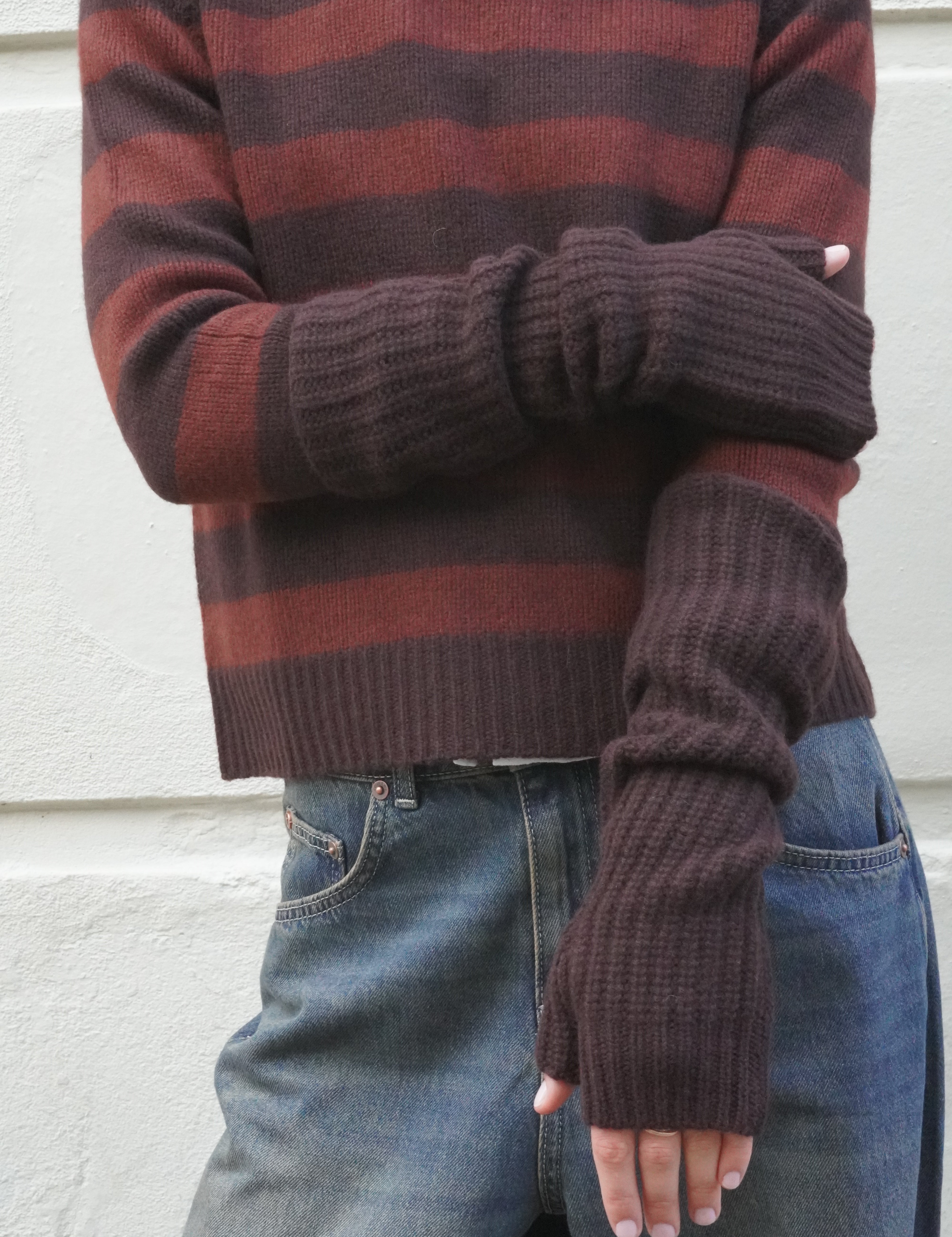 A person wearing a ribbed knit sweater with horizontal rust red and dark brown stripes, paired with blue jeans and matching dark brown fingerless knit gloves, standing against a light textured wall.