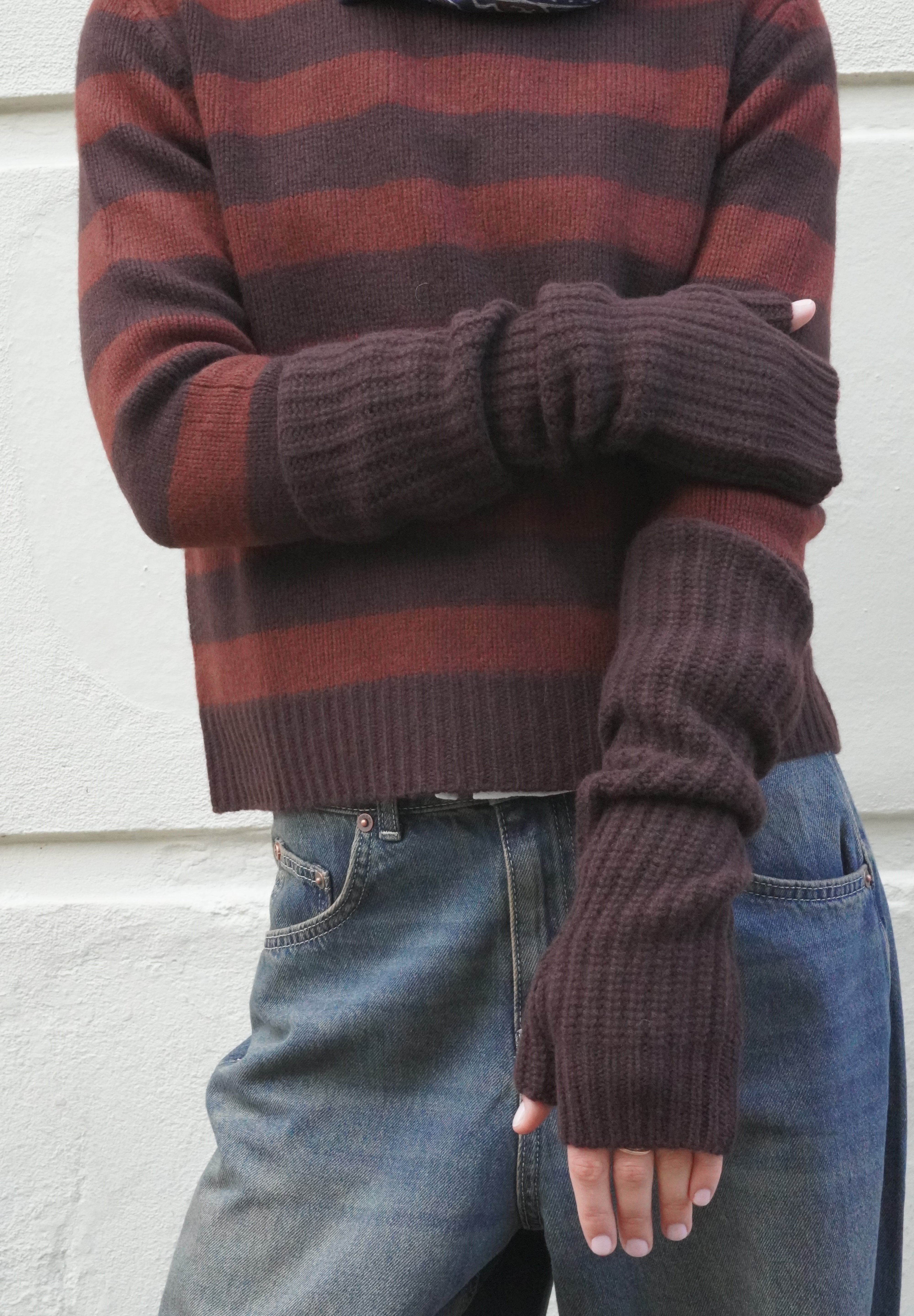A person wearing a ribbed knit sweater with horizontal rust red and dark brown stripes, paired with blue jeans and matching dark brown fingerless knit gloves, standing against a light textured wall.