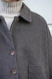 Close-up of a dark gray ribbed corduroy button-up shirt jacket worn over a light beige ribbed turtleneck sweater.
