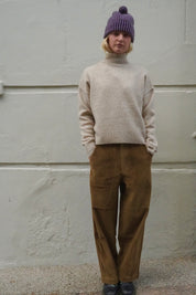 A person standing against a light textured wall wearing a cream turtleneck sweater, Olive Structured Corduroys, a purple knit beanie, and black shoes.