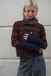 A woman wearing a dark brown and burgundy striped knit sweater, a navy blue paisley scarf, and matching navy blue fingerless knit sleeves, standing against a light-colored textured wall.