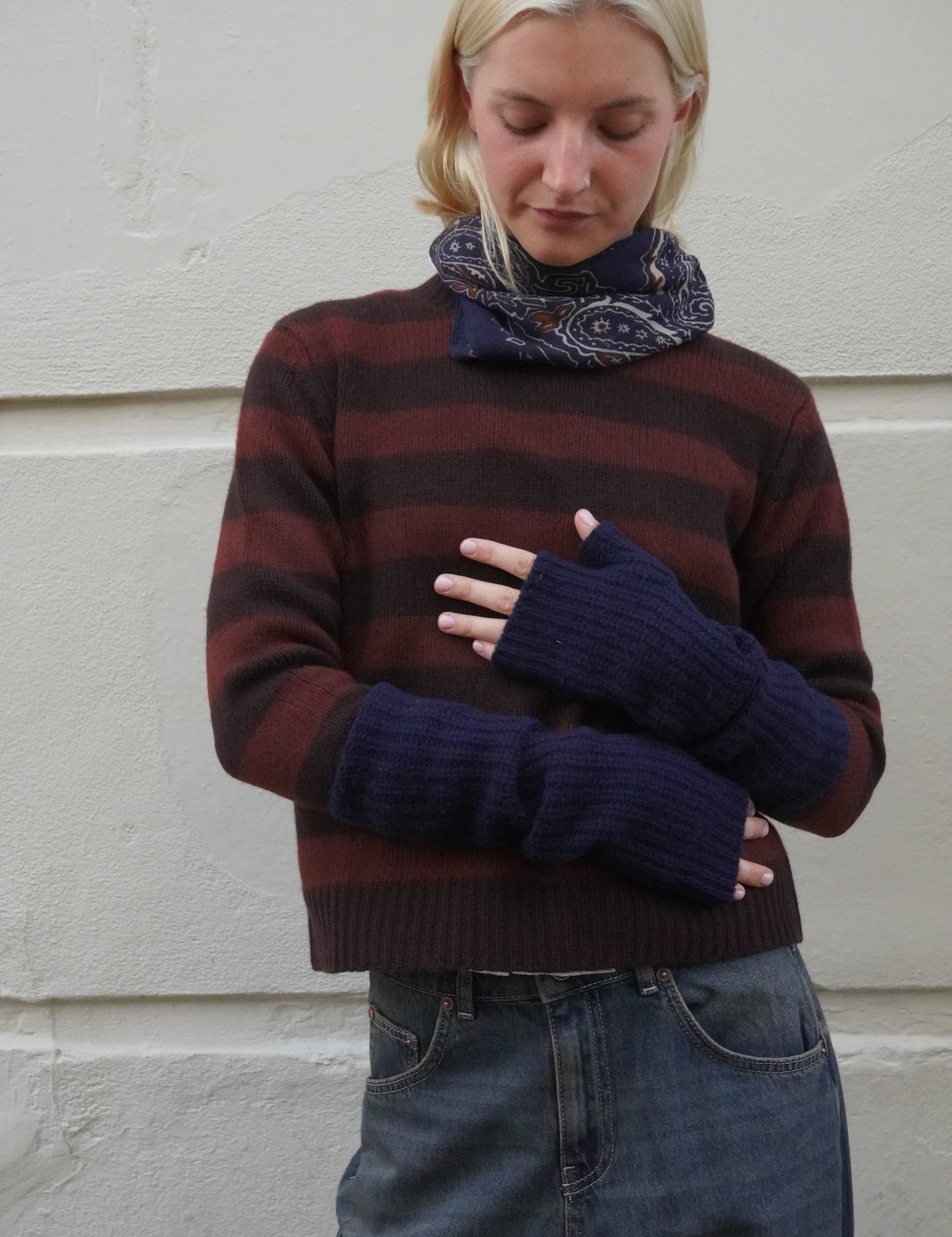 A woman wearing a dark brown and burgundy striped knit sweater, a navy blue paisley scarf, and matching navy blue fingerless knit sleeves, standing against a light-colored textured wall.