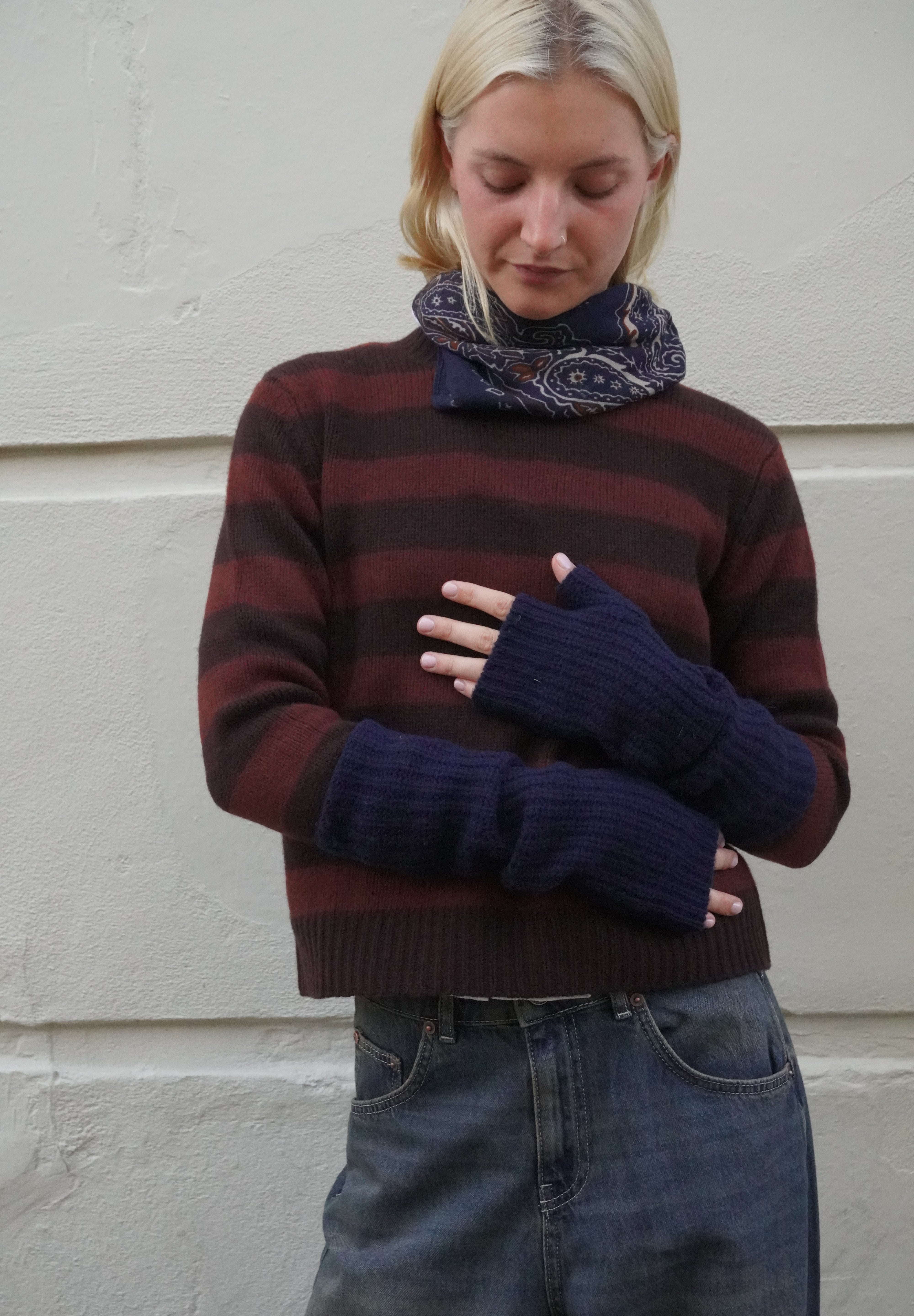 A woman wearing a dark brown and burgundy striped knit sweater, a navy blue paisley scarf, and matching navy blue fingerless knit sleeves, standing against a light-colored textured wall.
