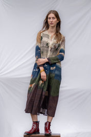 A woman stands against a white draped fabric backdrop wearing a long-sleeved, below knee-length dress with a multicolor print of blue, green, brown, and beige. 