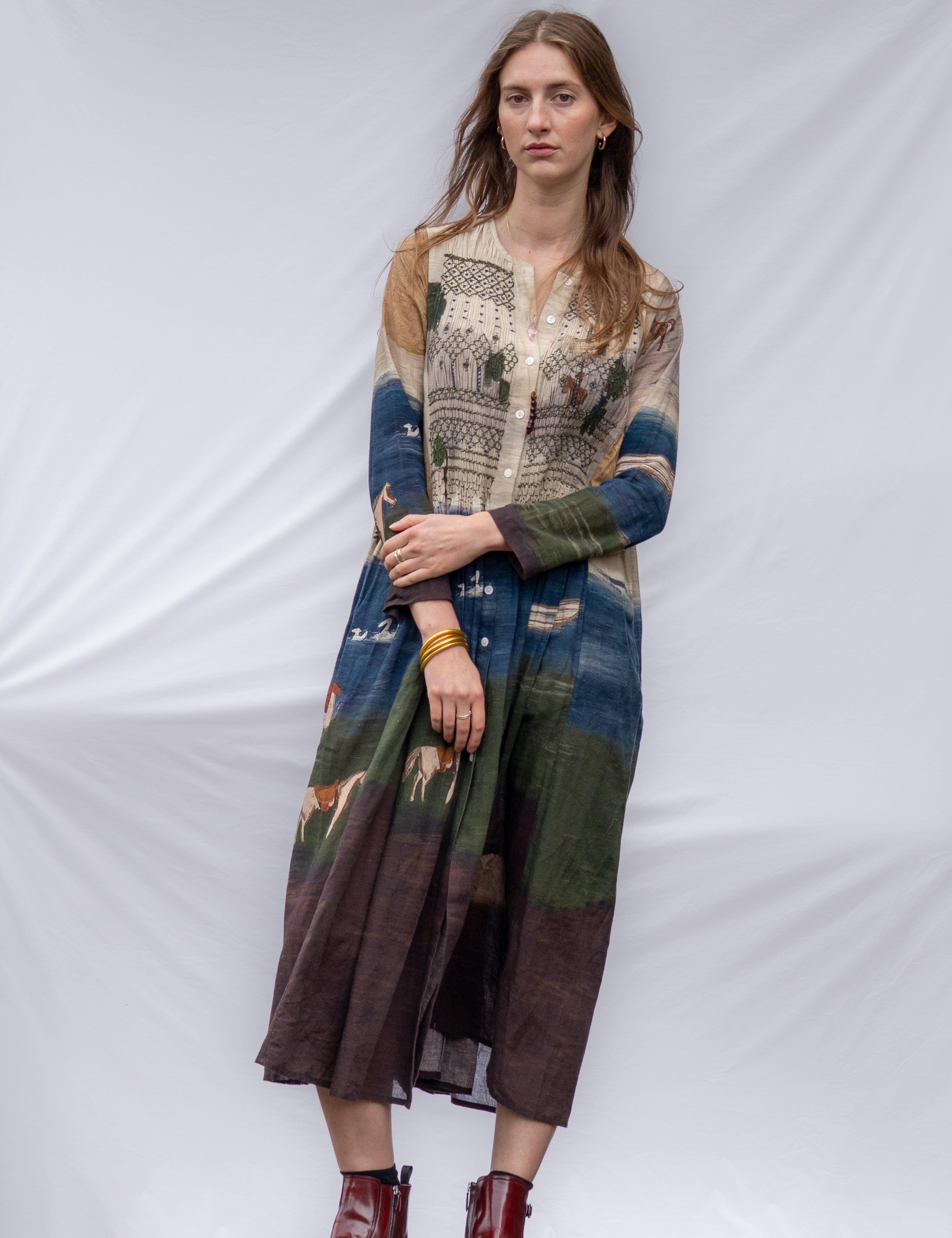 A woman stands against a white draped fabric backdrop wearing a long-sleeved, below knee-length dress with a multicolor print of blue, green, brown, and beige. 