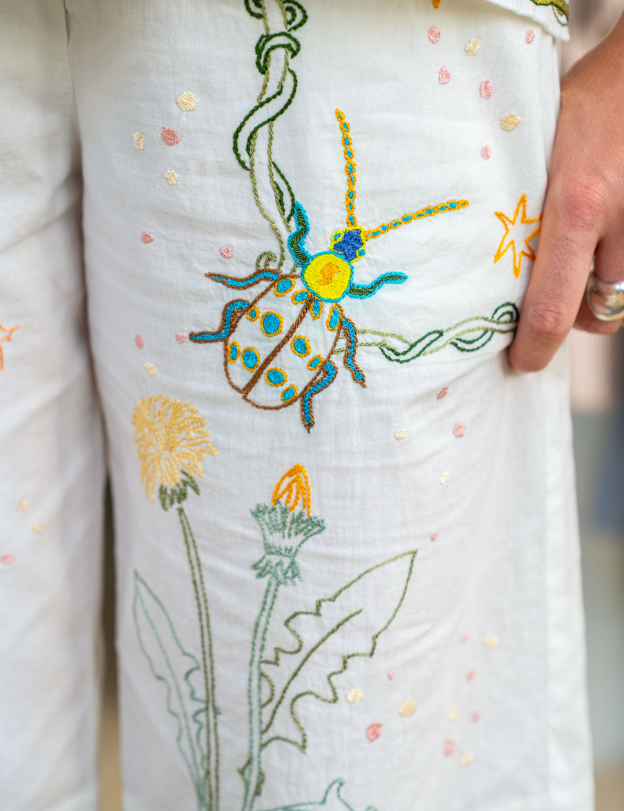 Close-up of a white embroidered pyjamas featuring a colourful beetle motif and green leafy stems, with a hand holding the fabric.