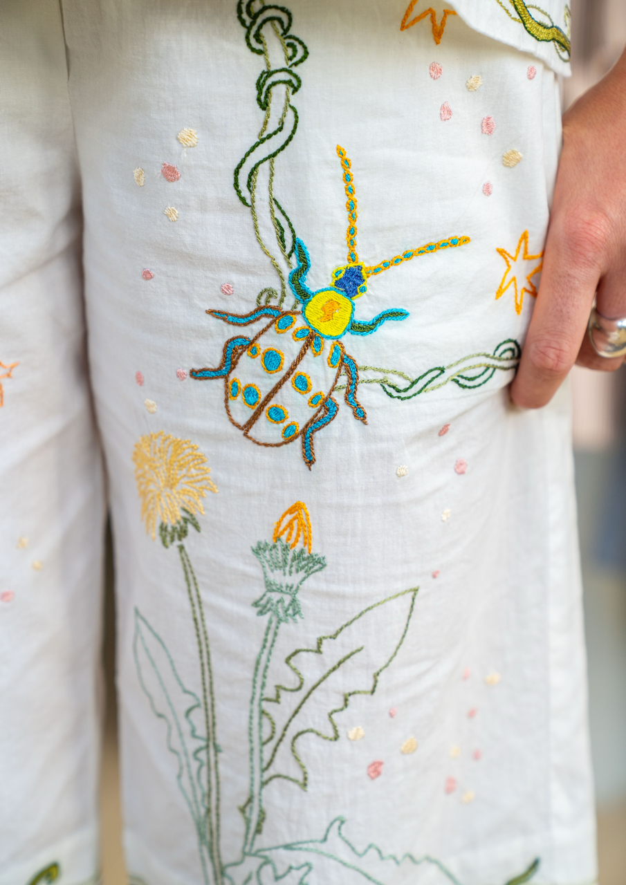 Close-up of a white embroidered pyjamas featuring a colourful beetle motif and green leafy stems, with a hand holding the fabric.