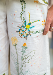 Close-up of a white embroidered pyjamas featuring a colourful beetle motif and green leafy stems, with a hand holding the fabric.