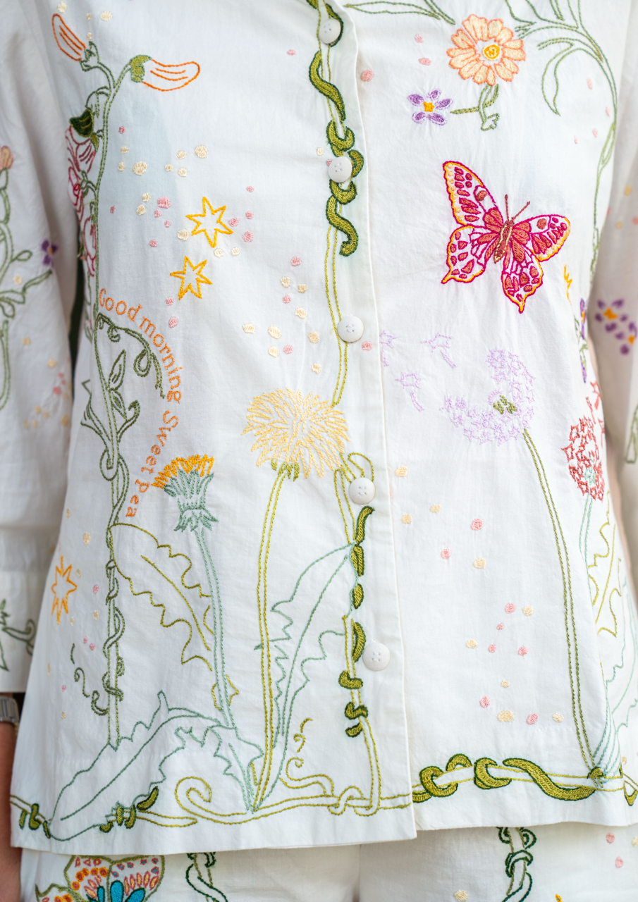 Close-up of a white embroidered pyjama top with colorful floral and butterfly motifs, including green vines, yellow stars, pink flowers, and a pink butterfly on the right side.