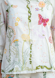 Close-up of a white embroidered pyjama top with colorful floral and butterfly motifs, including green vines, yellow stars, pink flowers, and a pink butterfly on the right side.