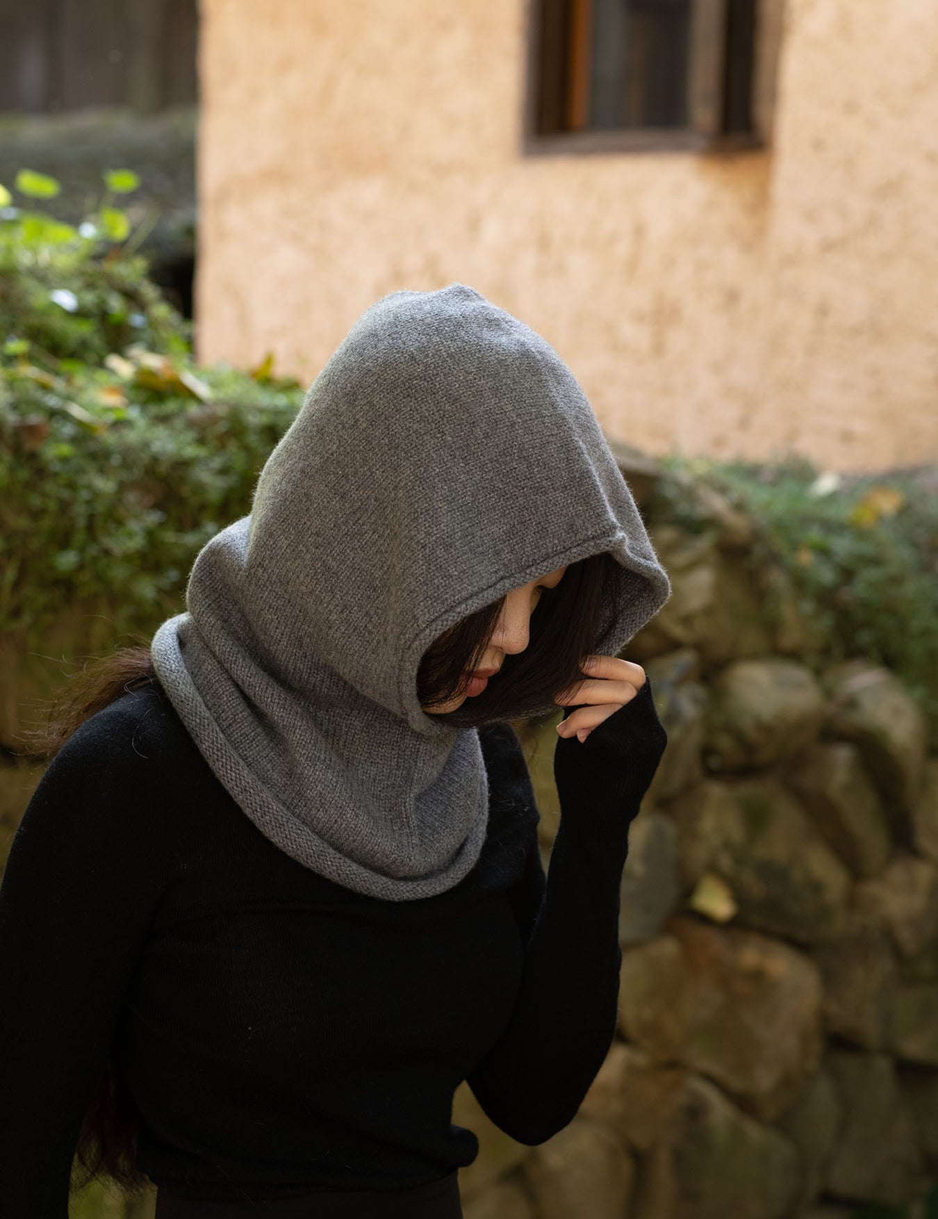 A person wearing a gray knit hooded neck warmer or balaclava-style hood over a black top, standing outdoors in front of a stone wall and beige building wall.