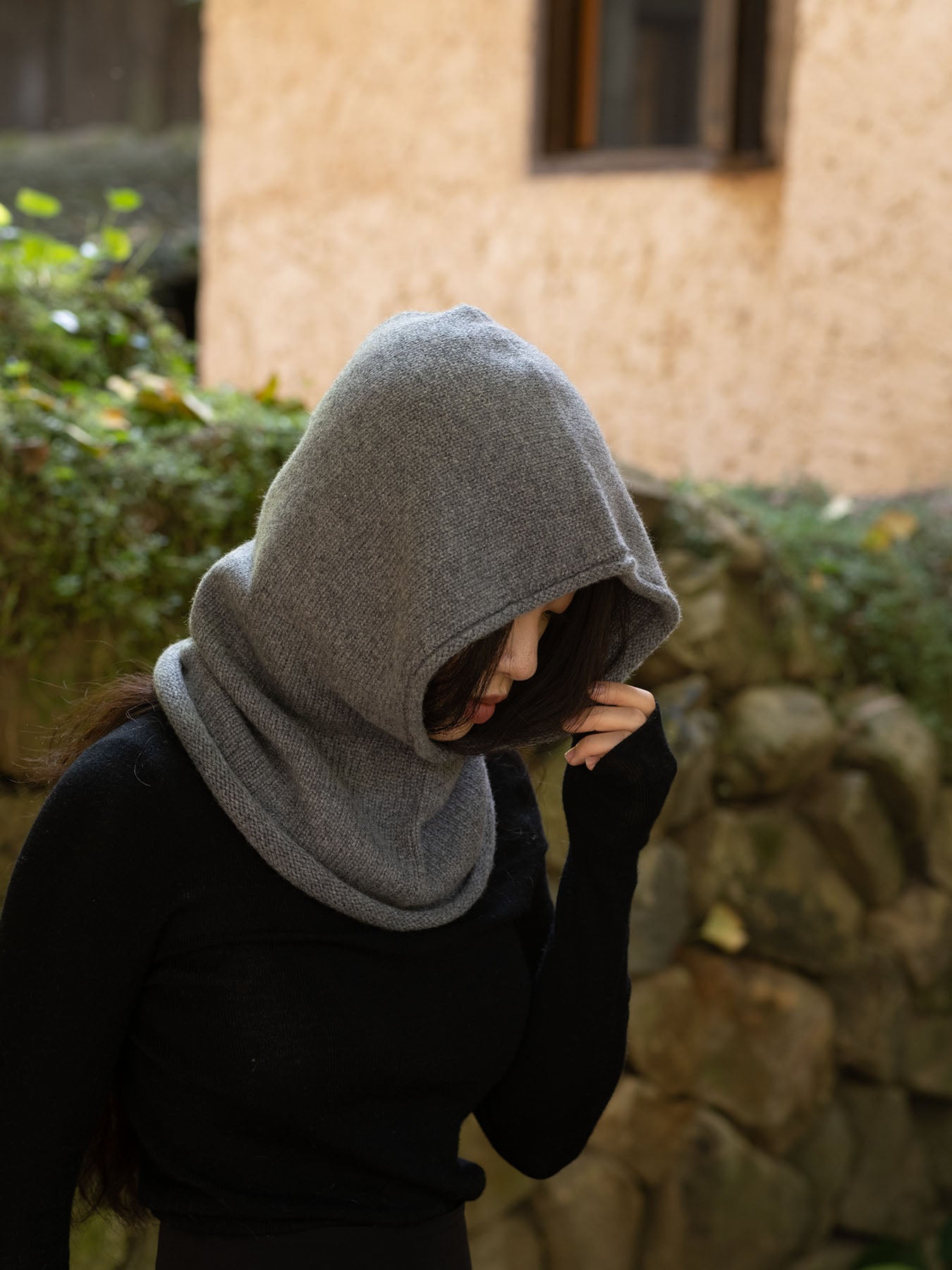A person wearing a gray knit hooded neck warmer or balaclava-style hood over a black top, standing outdoors in front of a stone wall and beige building wall.