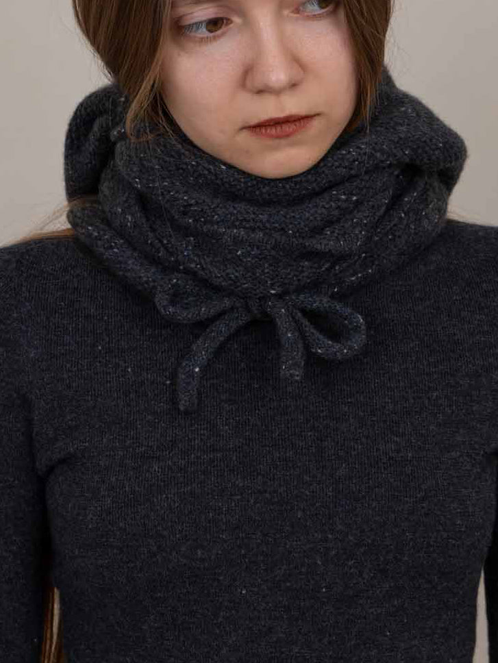 A woman wearing a dark charcoal gray knit cowl neck warmer with a bow detail, styled over a dark gray long-sleeve top against a plain beige background.