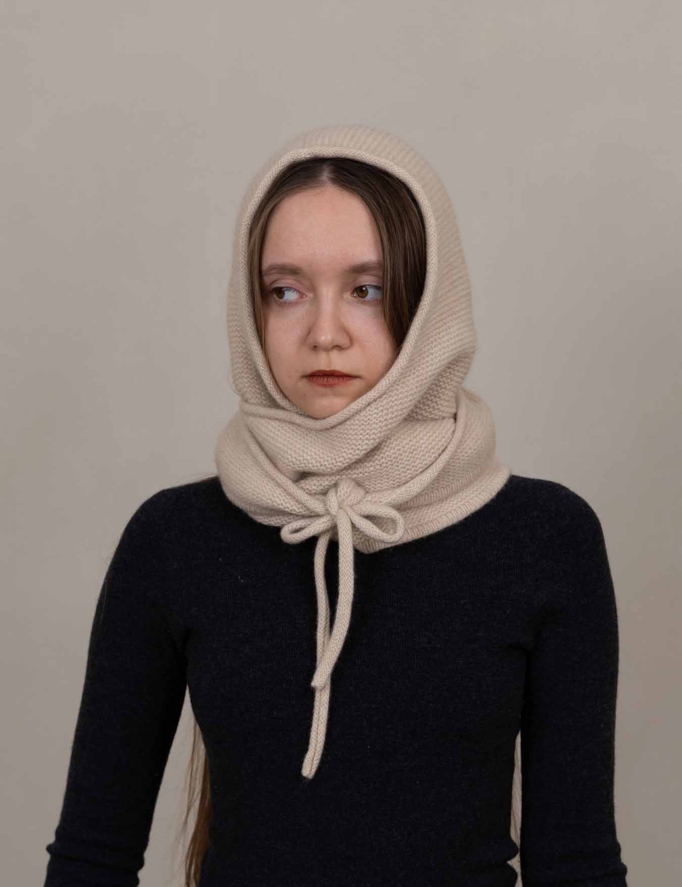 A woman wearing a cream-colored knit head and neck scarf tied at the front, paired with a dark navy long-sleeve top, against a plain light gray background.