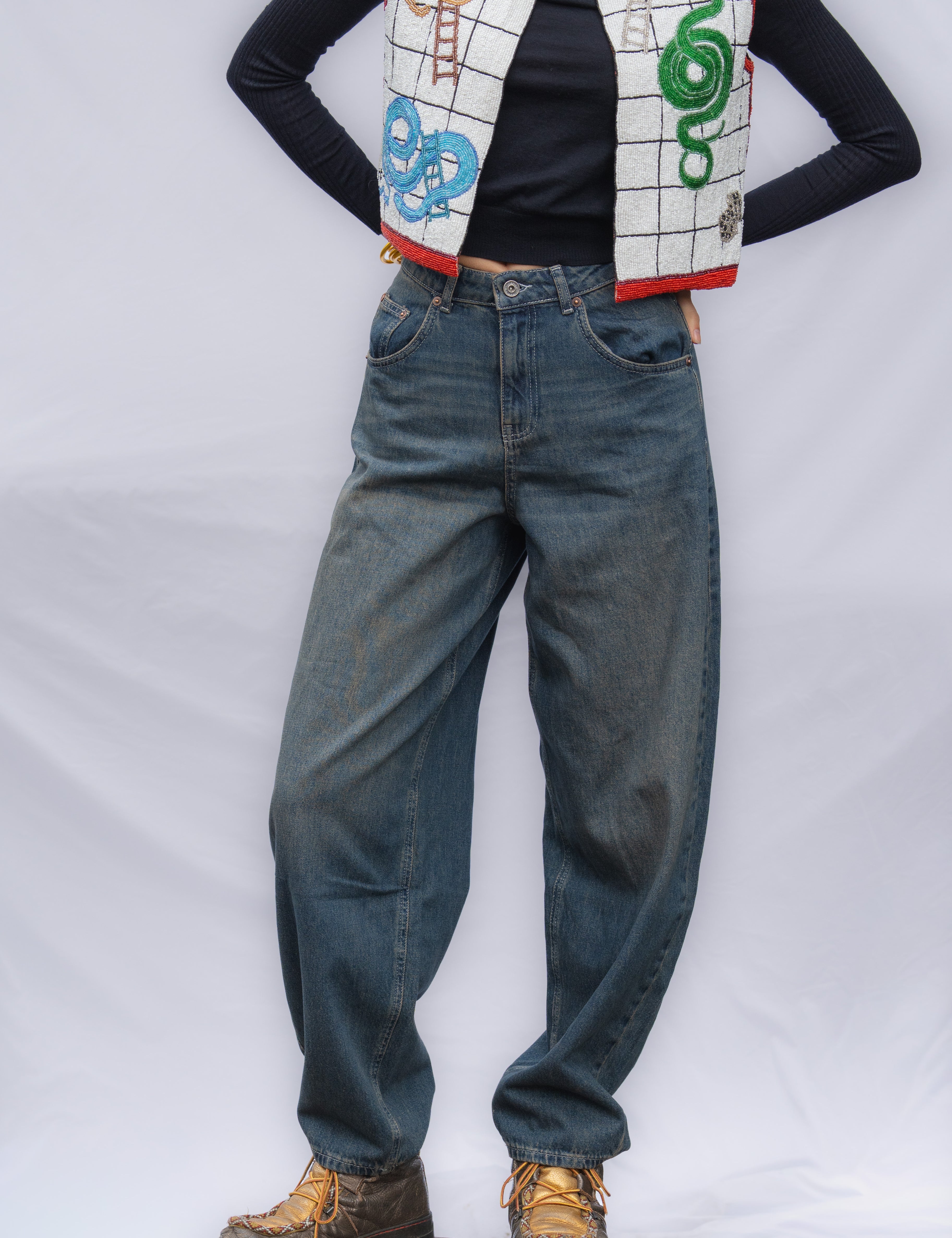 A person wearing loose-fit blue jeans, a black long-sleeve top, a white beaded vest with snakes and ladder designs,, standing on a wooden block.