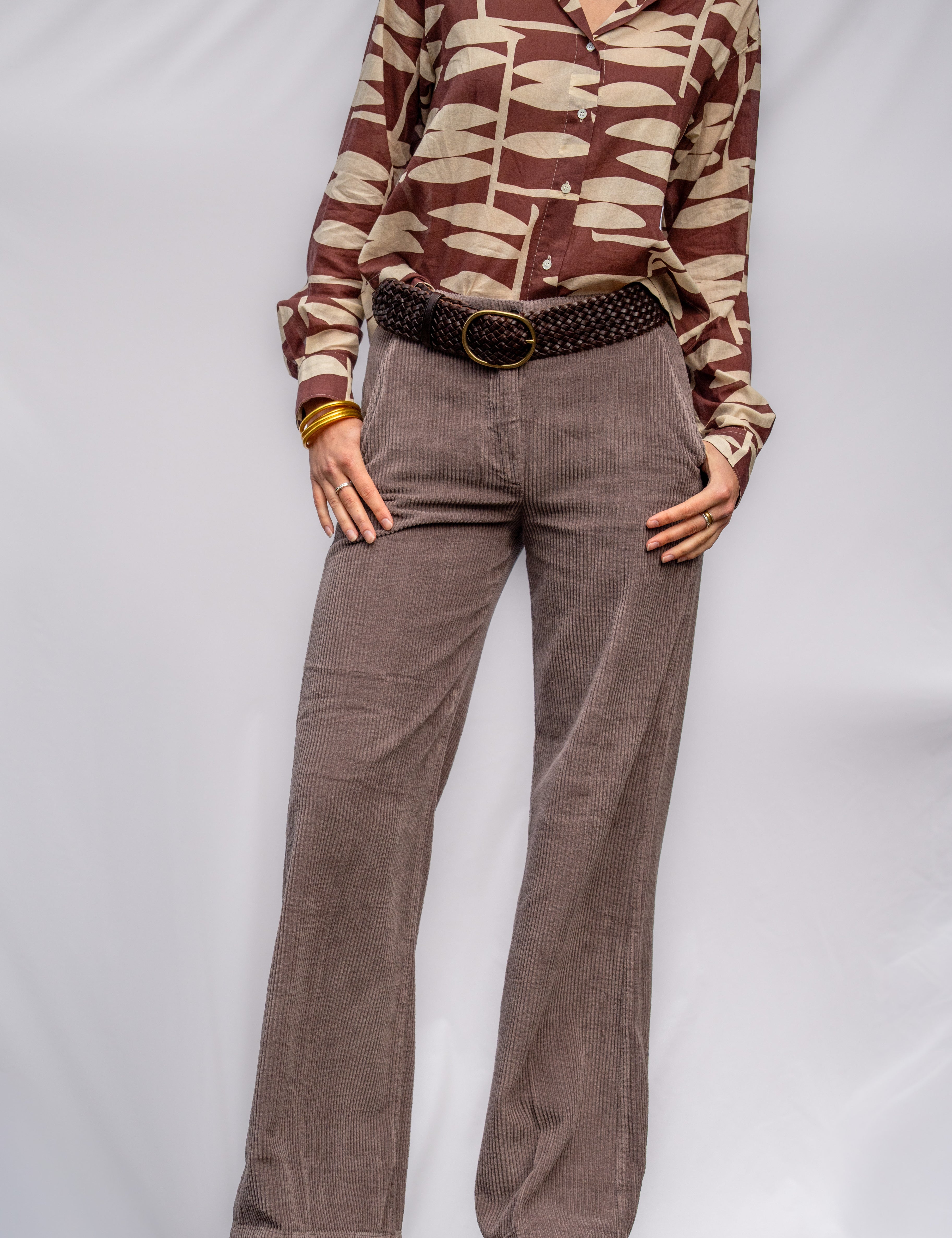 A model wearing a brown clove shirt, taupe wide-leg trousers, a brown woven leather belt, standing against a white studio backdrop.