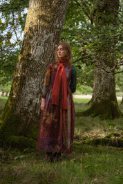 A woman stands in a mossy forest next to a large tree trunk, wearing a long bohemian-style dress with a rust-red scarf. 