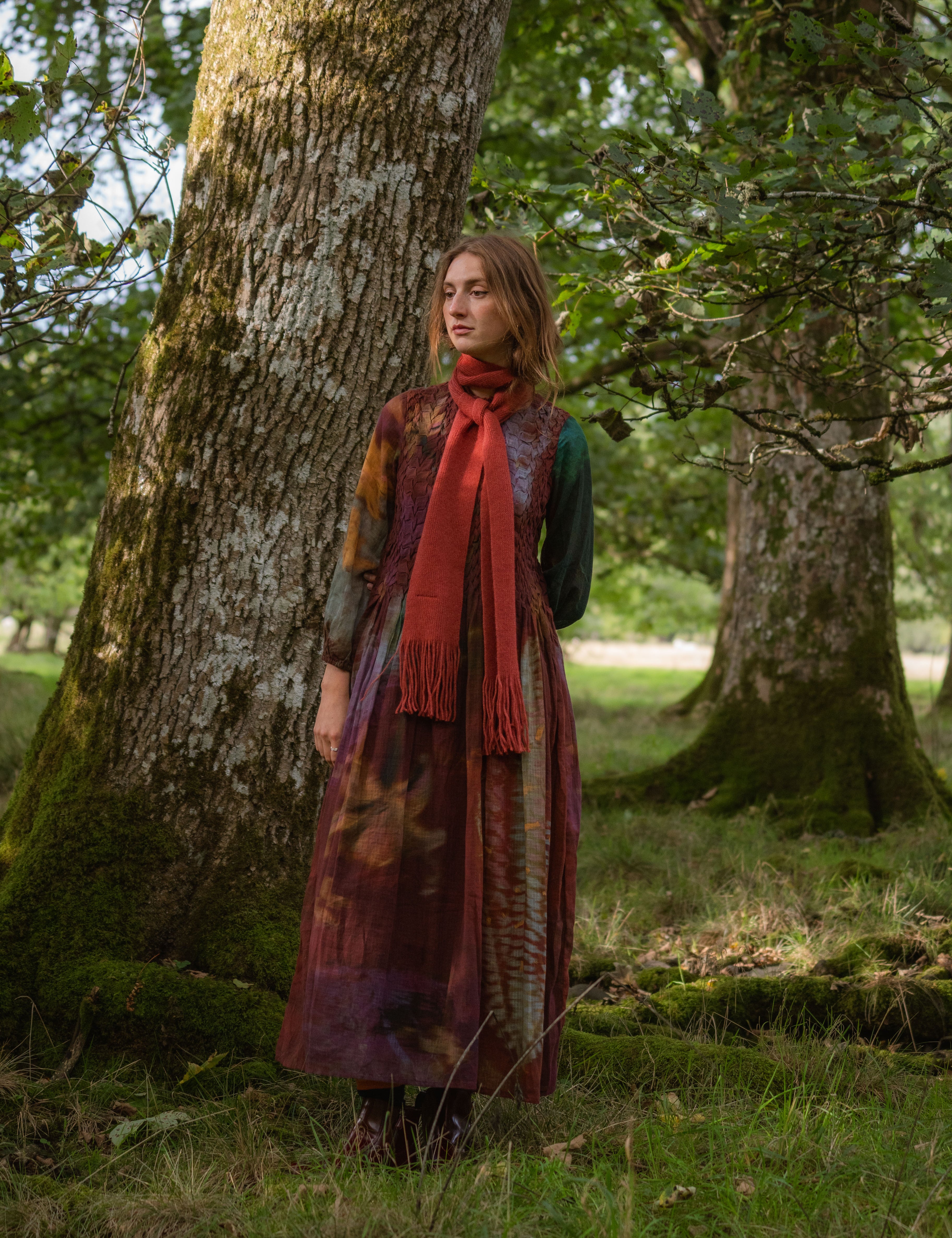 A woman stands in a mossy forest next to a large tree trunk, wearing a long bohemian-style dress with a rust-red scarf. 