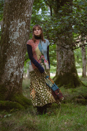A woman stands in forest, wearing a long flowy patterned dress, that has a painting like print. 