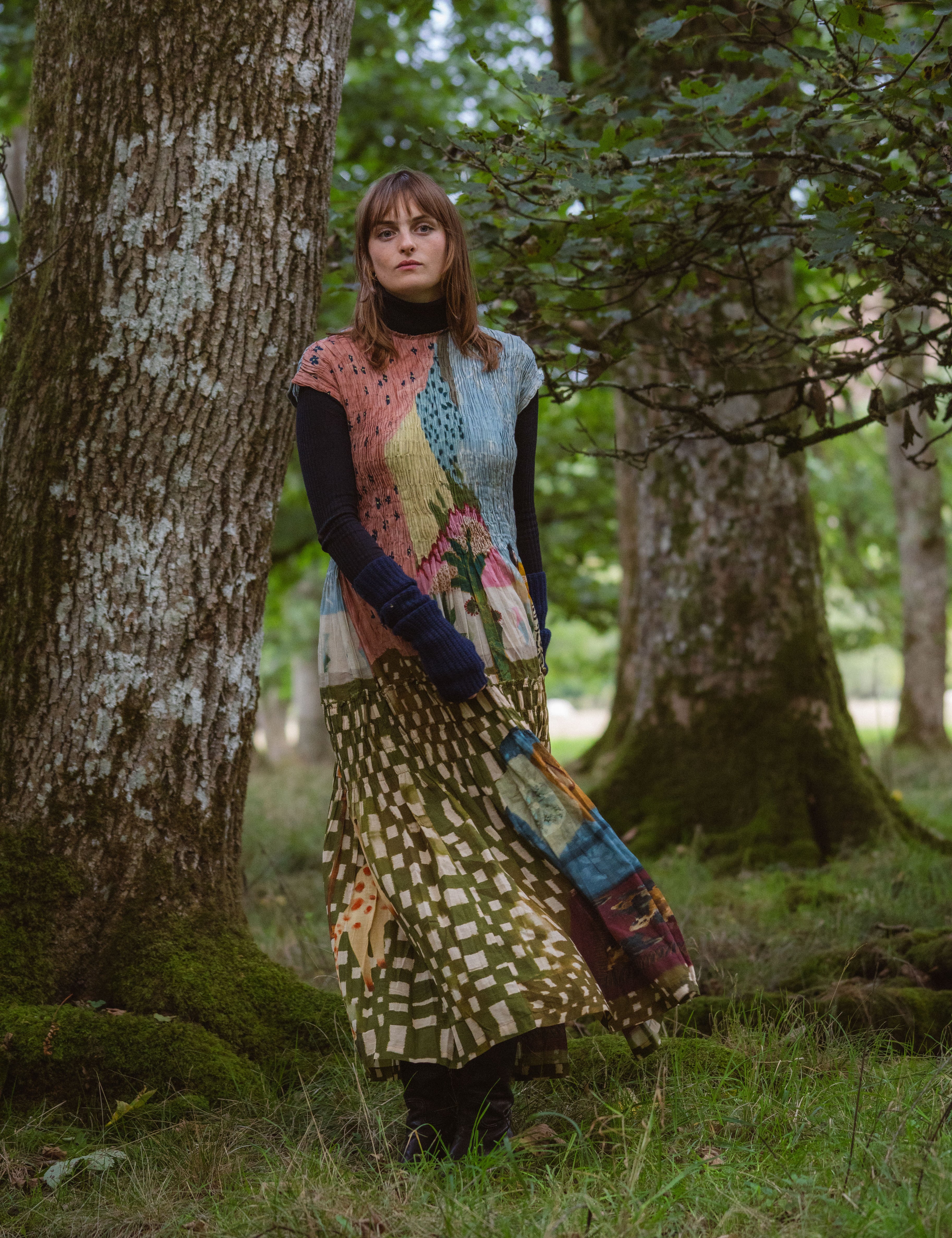 A woman stands in forest, wearing a long flowy patterned dress, that has a painting like print. 