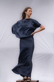 A woman modeling a navy long, flowy dress. The dress is ankle length and has one longer draping sleeve and the left sleeve is shorter. 