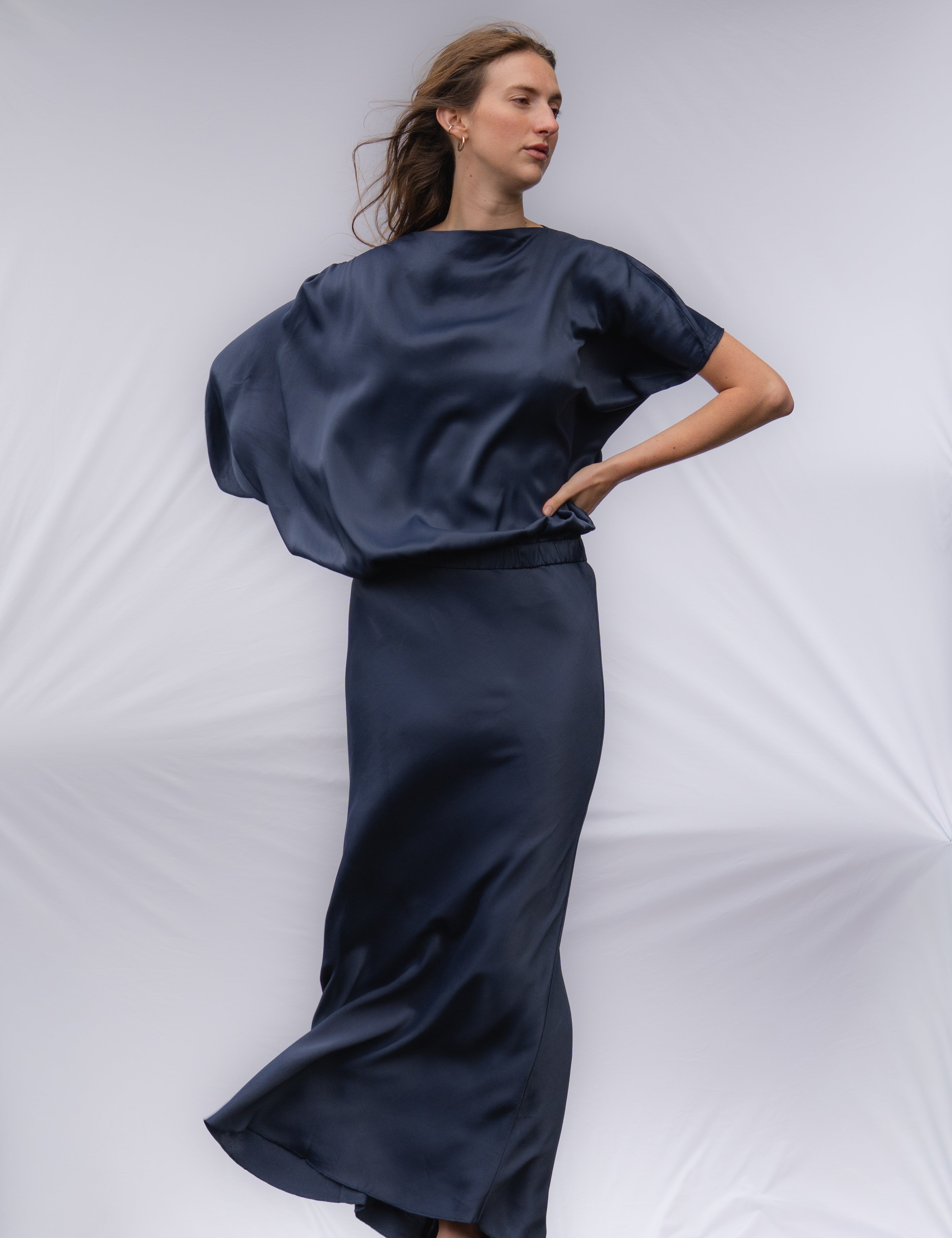 A woman modeling a navy long, flowy dress. The dress is ankle length and has one longer draping sleeve and the left sleeve is shorter. 