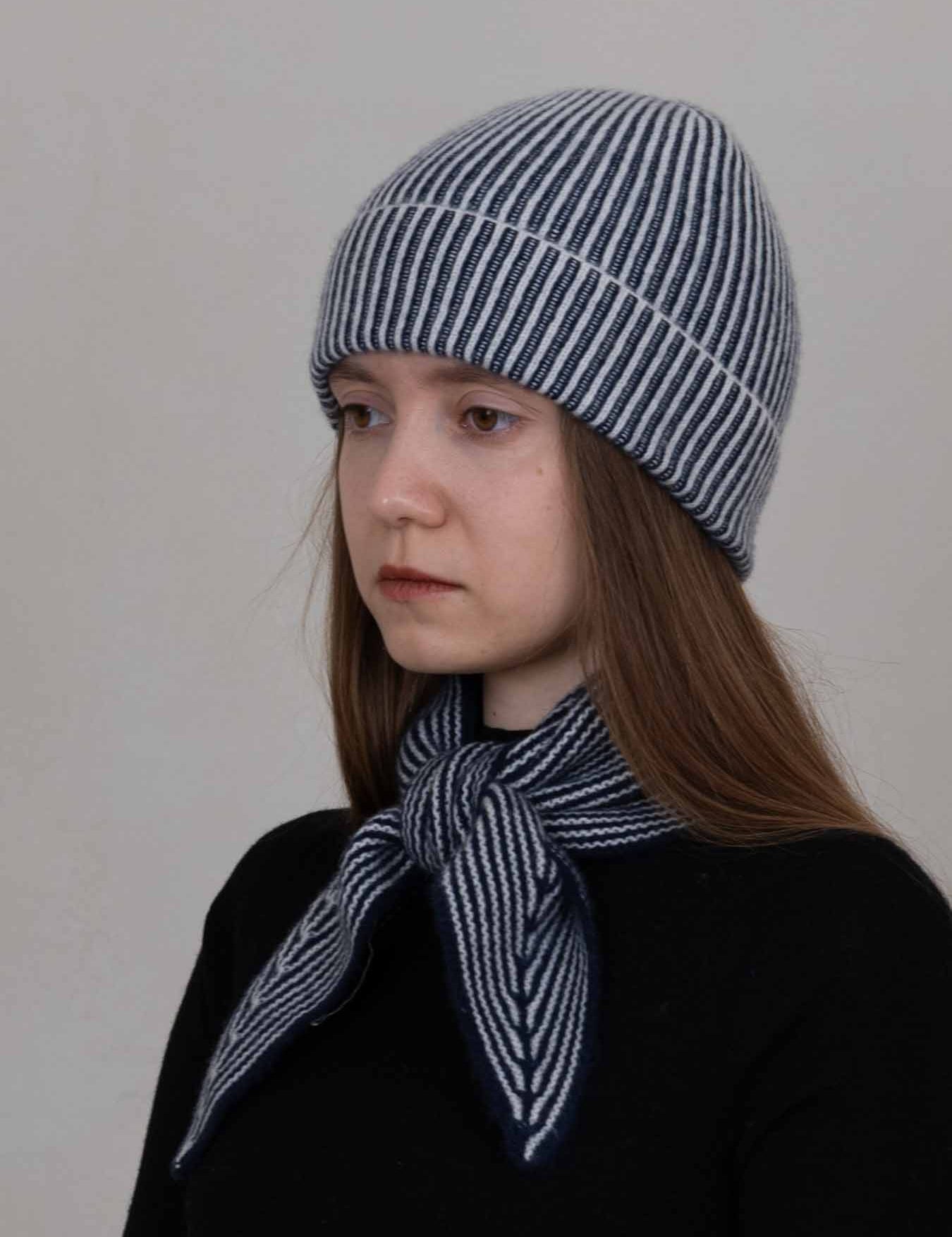 A woman wearing a navy and white striped knit beanie and a matching striped neck scarf, photographed against a plain light gray background.
