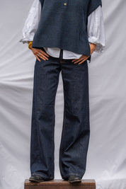 A person wearing dark blue wide-leg denim jeans, a white long-sleeve blouse, and a dark blue knit vest, standing on a wooden block against a white draped backdrop.