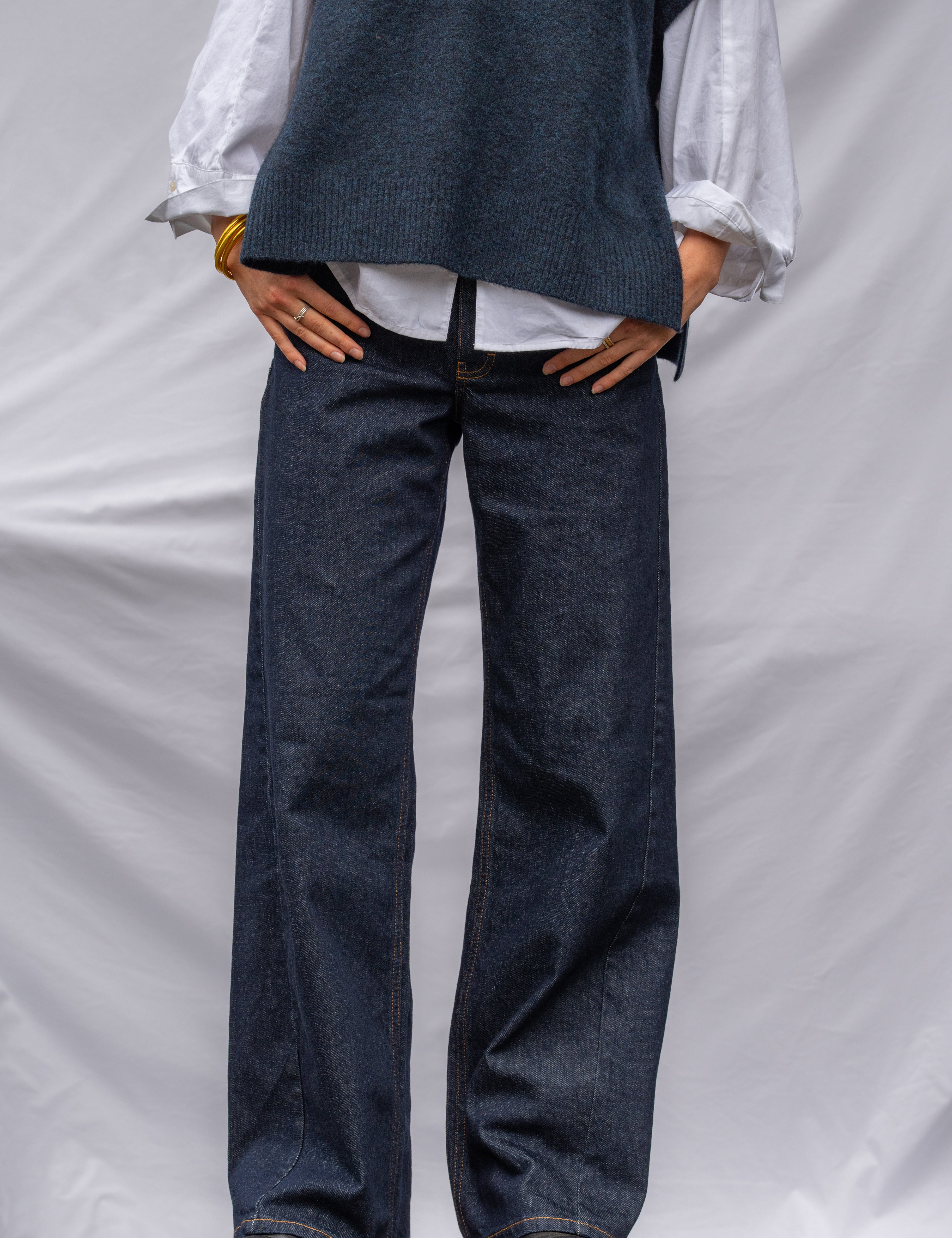 A person wearing dark blue wide-leg denim jeans, a white long-sleeve blouse, and a dark blue knit vest, standing on a wooden block against a white draped backdrop.
