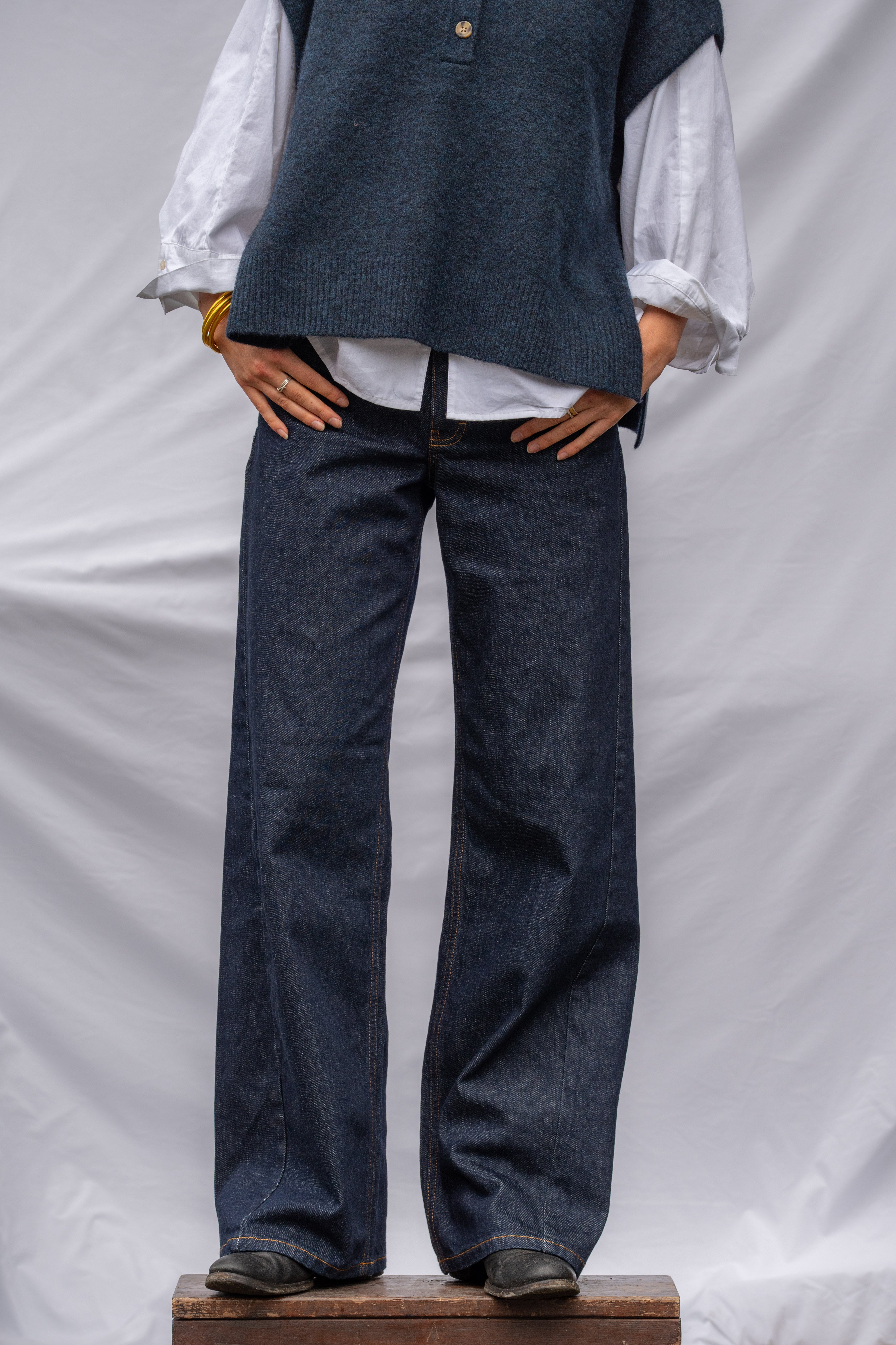A person wearing dark blue wide-leg denim jeans, a white long-sleeve blouse, and a dark blue knit vest, standing on a wooden block against a white draped backdrop.