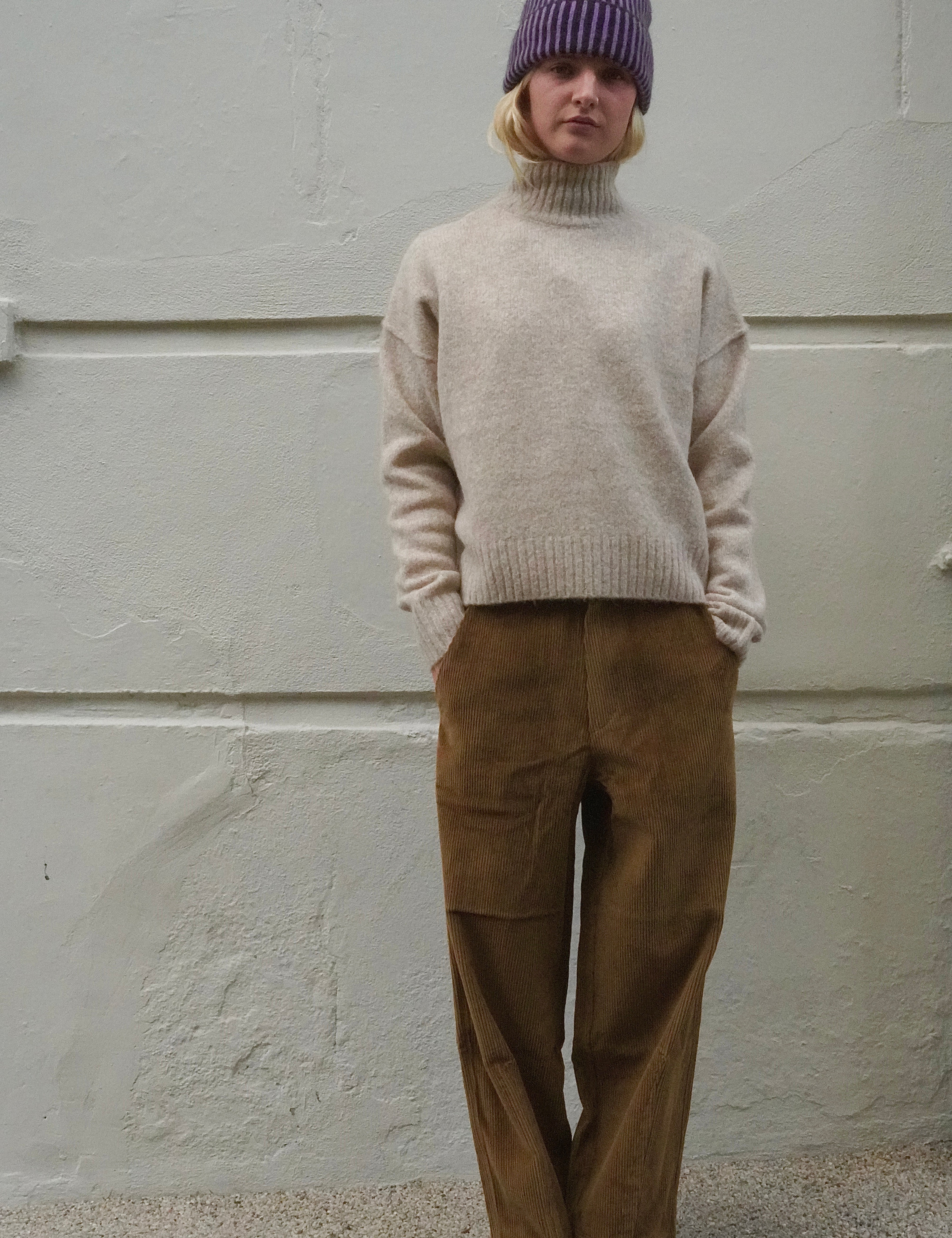 A person standing against a light textured wall wearing a cream turtleneck sweater, brown wide-leg pants, a purple knit beanie, and black shoes.