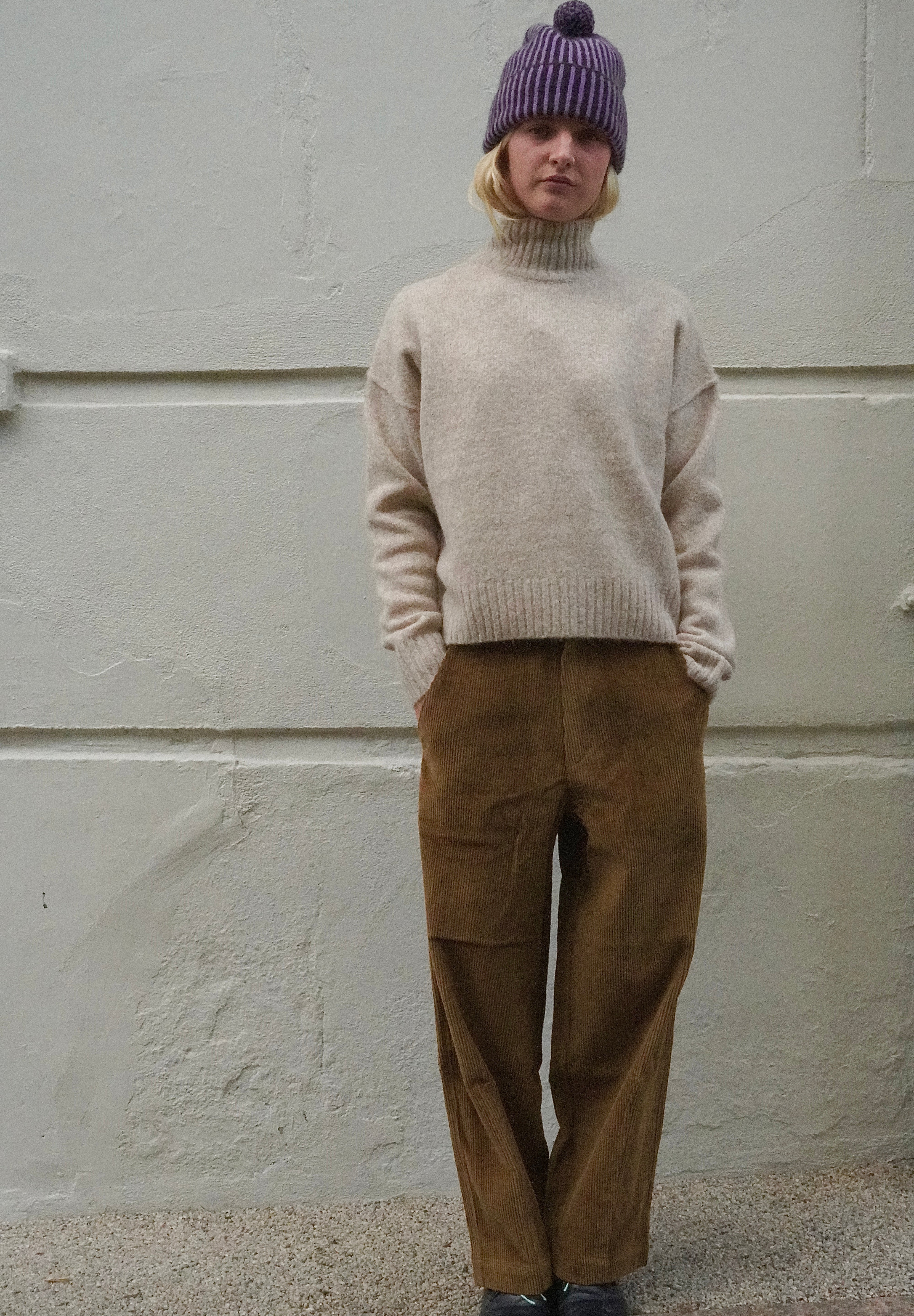 A person standing against a light textured wall wearing a cream turtleneck sweater, brown wide-leg pants, a purple knit beanie, and black shoes.
