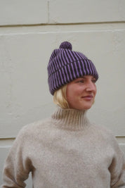 Woman wearing striped purple beanie with bobble on top, and a knitted cream polar neck jumper. 