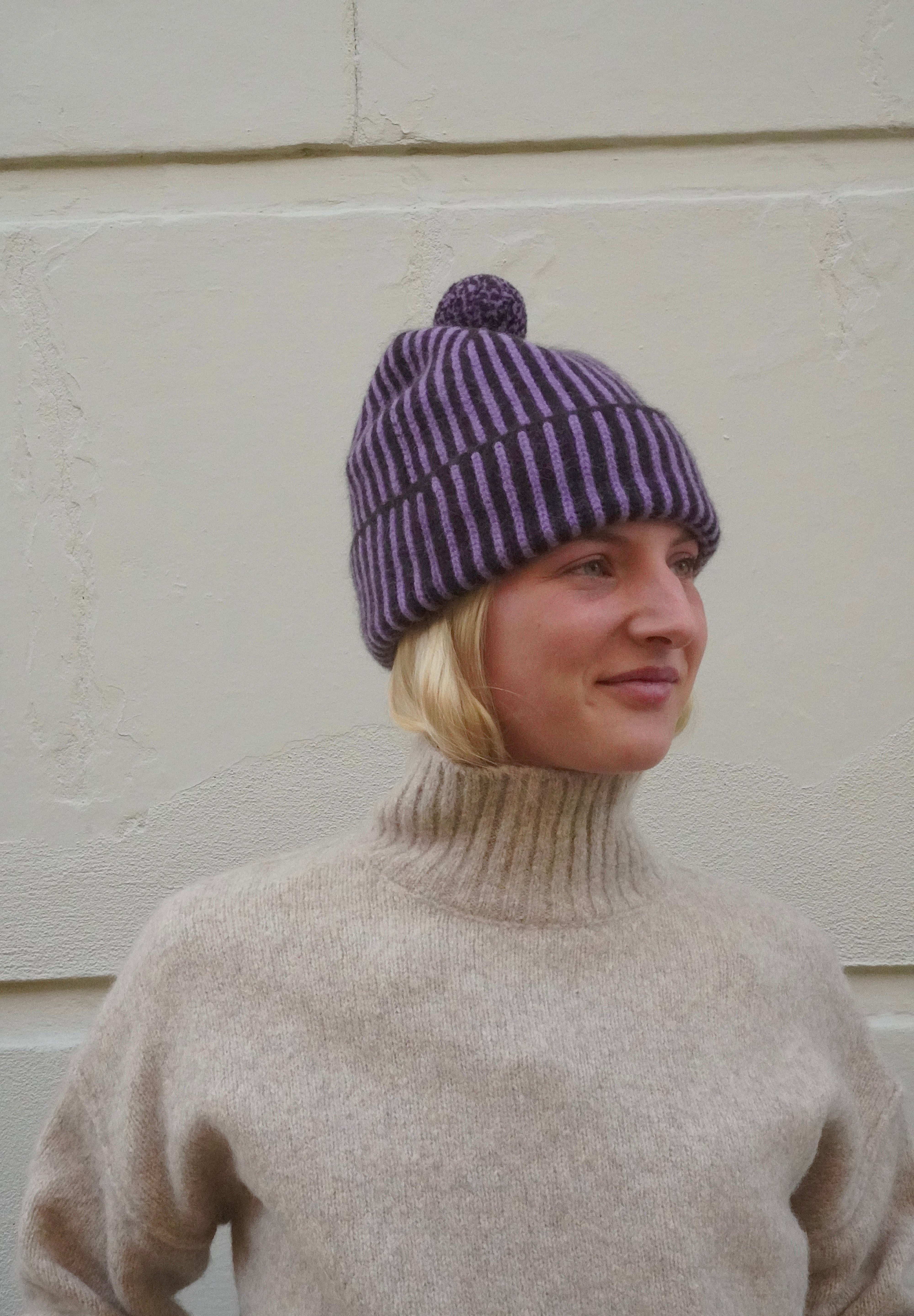 Woman wearing striped purple beanie with bobble on top, and a knitted cream polar neck jumper. 