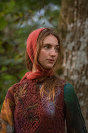 A woman outdoors wearing a rust-red head covering, standing in front of a tree trunk with blurred green foliage in the background.