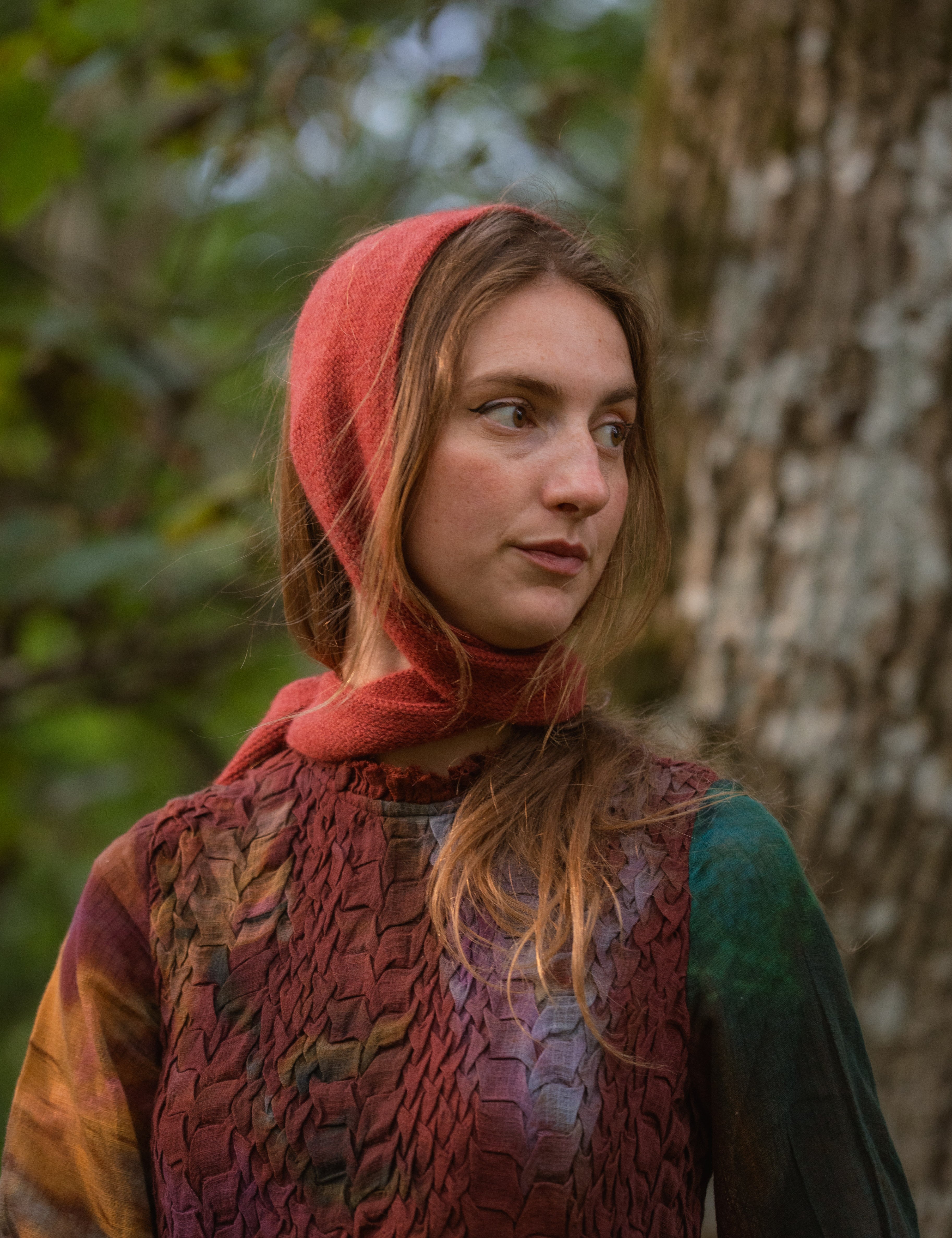 A woman outdoors wearing a rust-red head covering, standing in front of a tree trunk with blurred green foliage in the background.