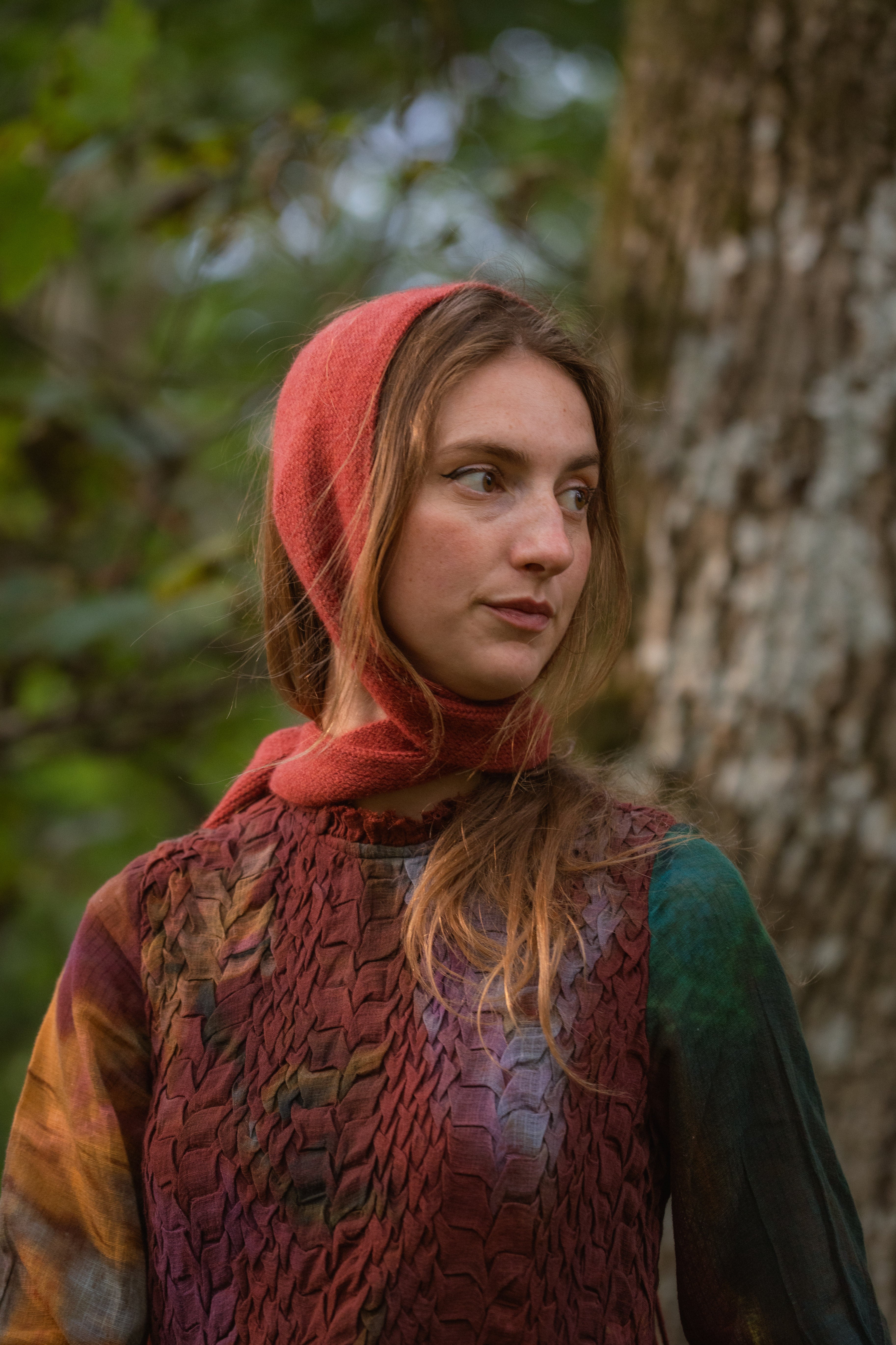 A woman outdoors wearing a rust-red head covering, standing in front of a tree trunk with blurred green foliage in the background.