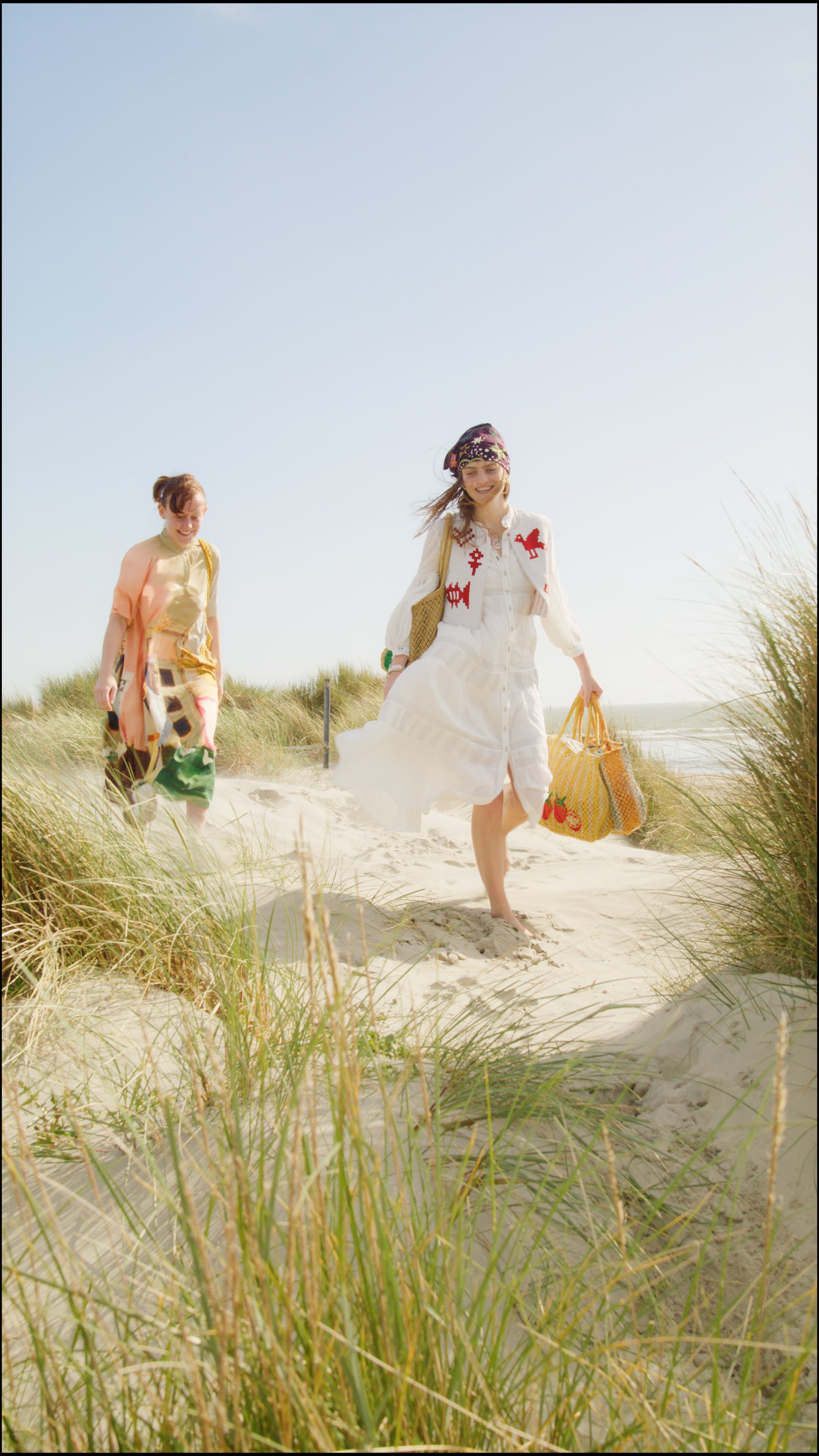 Two women walking on a sandy path with grass, one in a white dress and the other in a colorful outfit, with a clear blue sky.