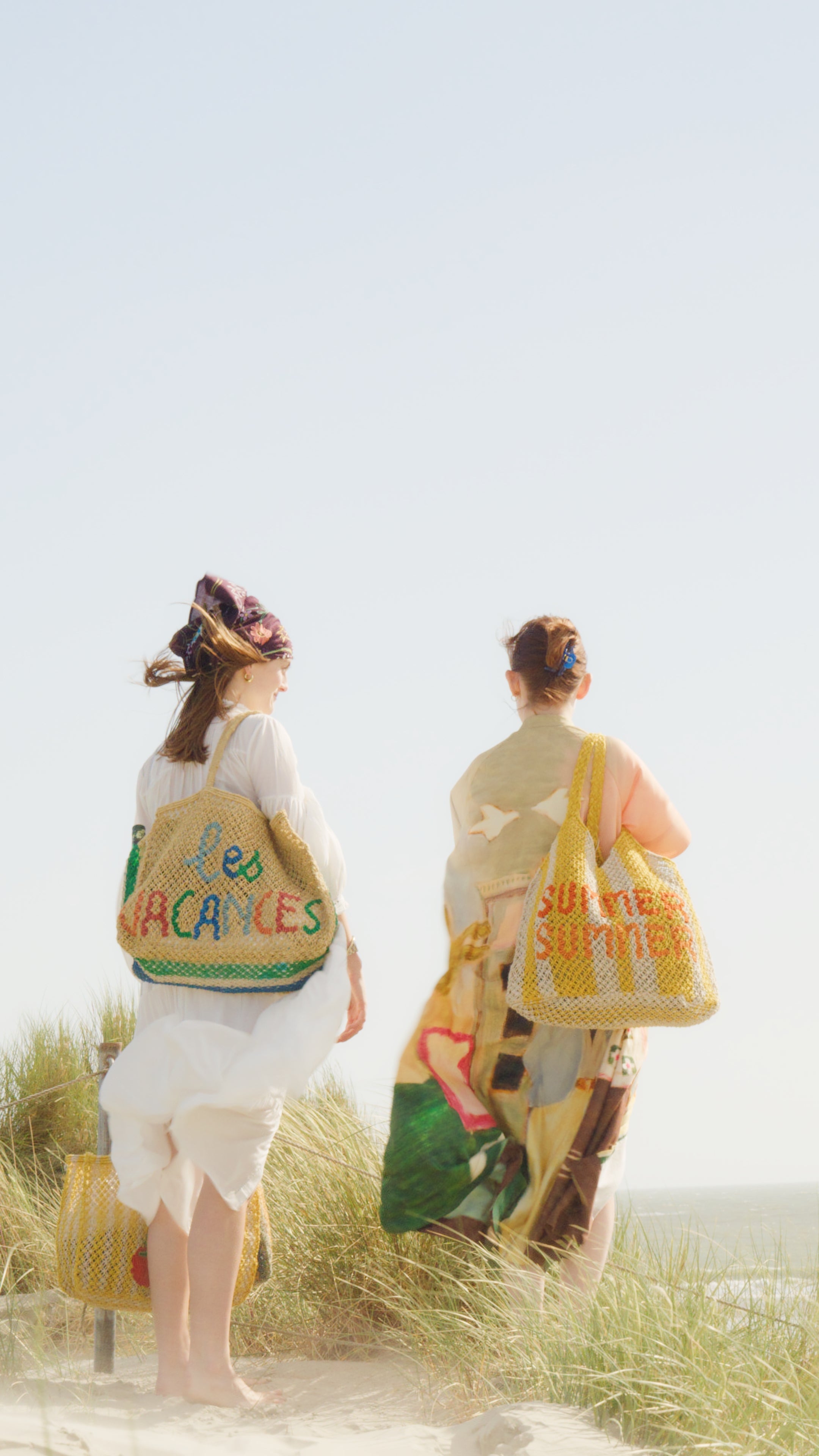 Two people on a beach with colourful jute bags, one labeled 'les vacances' and the other 'summer'.