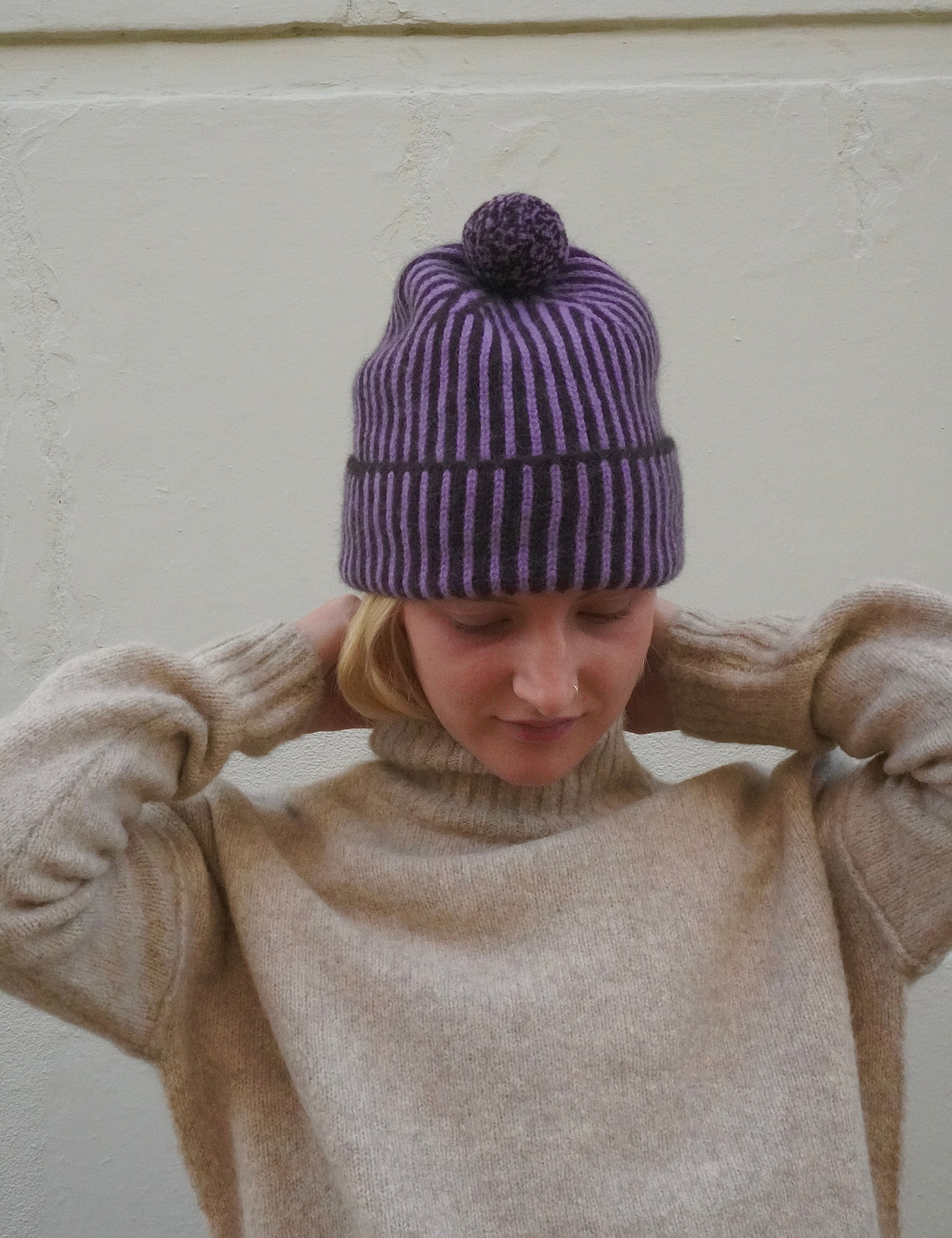 Woman wearing striped purple beanie with bobble on top, and a knitted cream polar neck jumper. 